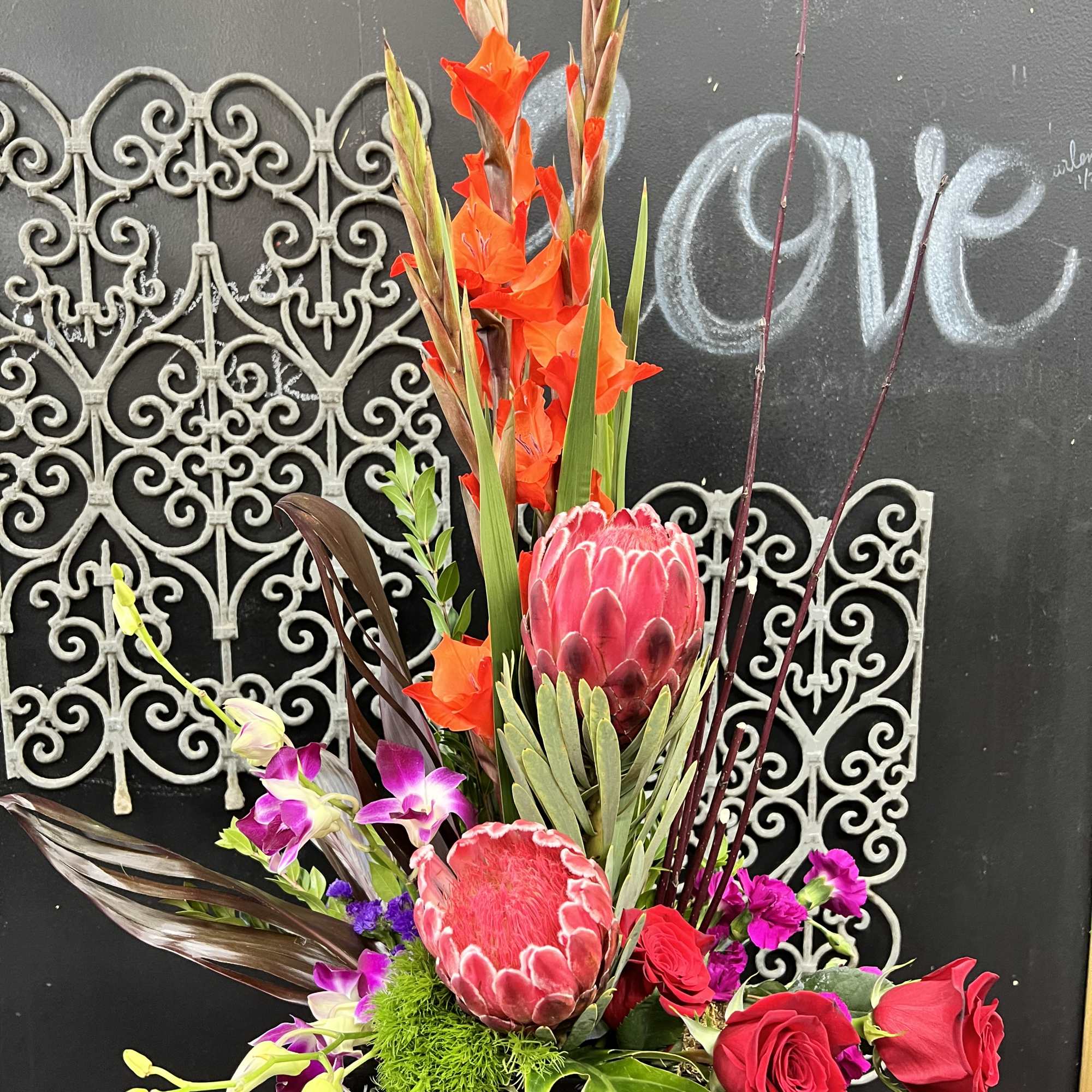 Tall tropical arrangement with red protea, roses, and orange blooms in a black vase