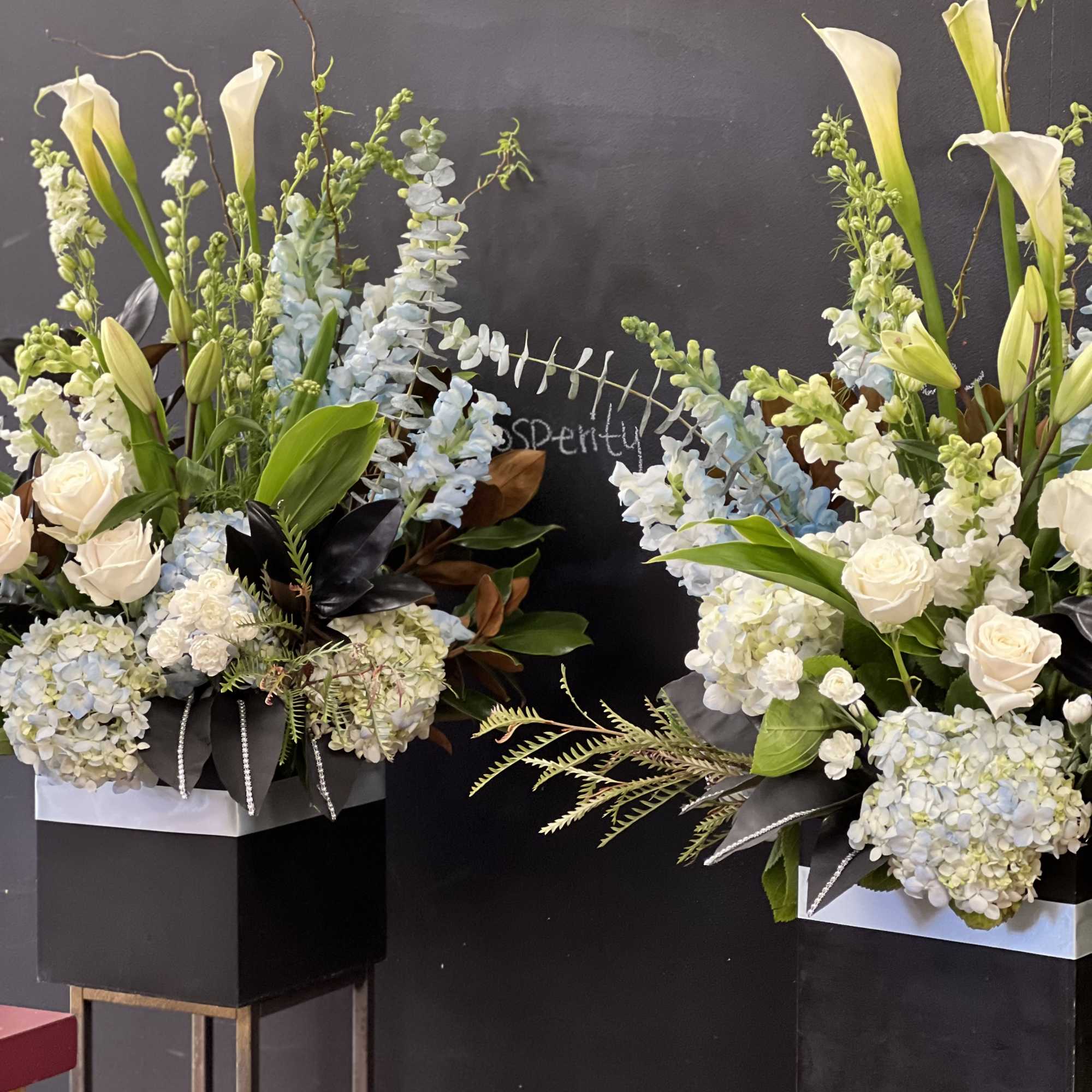 Two tall floral arrangements with white calla lilies, roses, and blue hydrangeas in black boxes