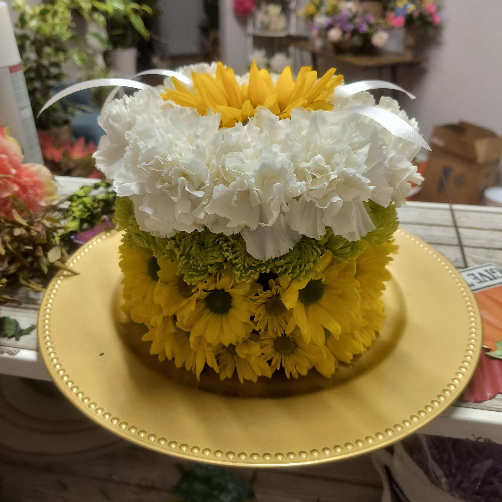 Round floral arrangement with yellow, white, and green flowers on a gold plate