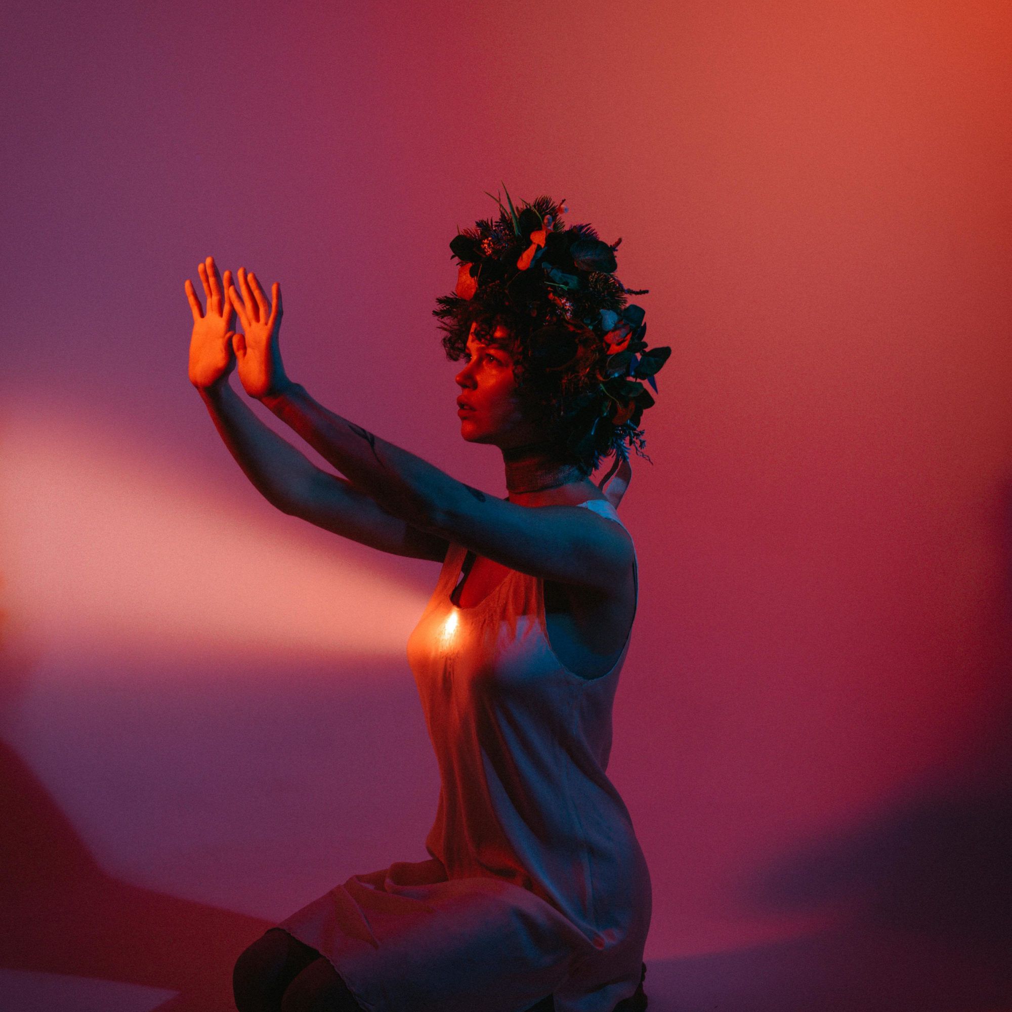 Person kneeling with a floral crown and raised hands under colored lighting