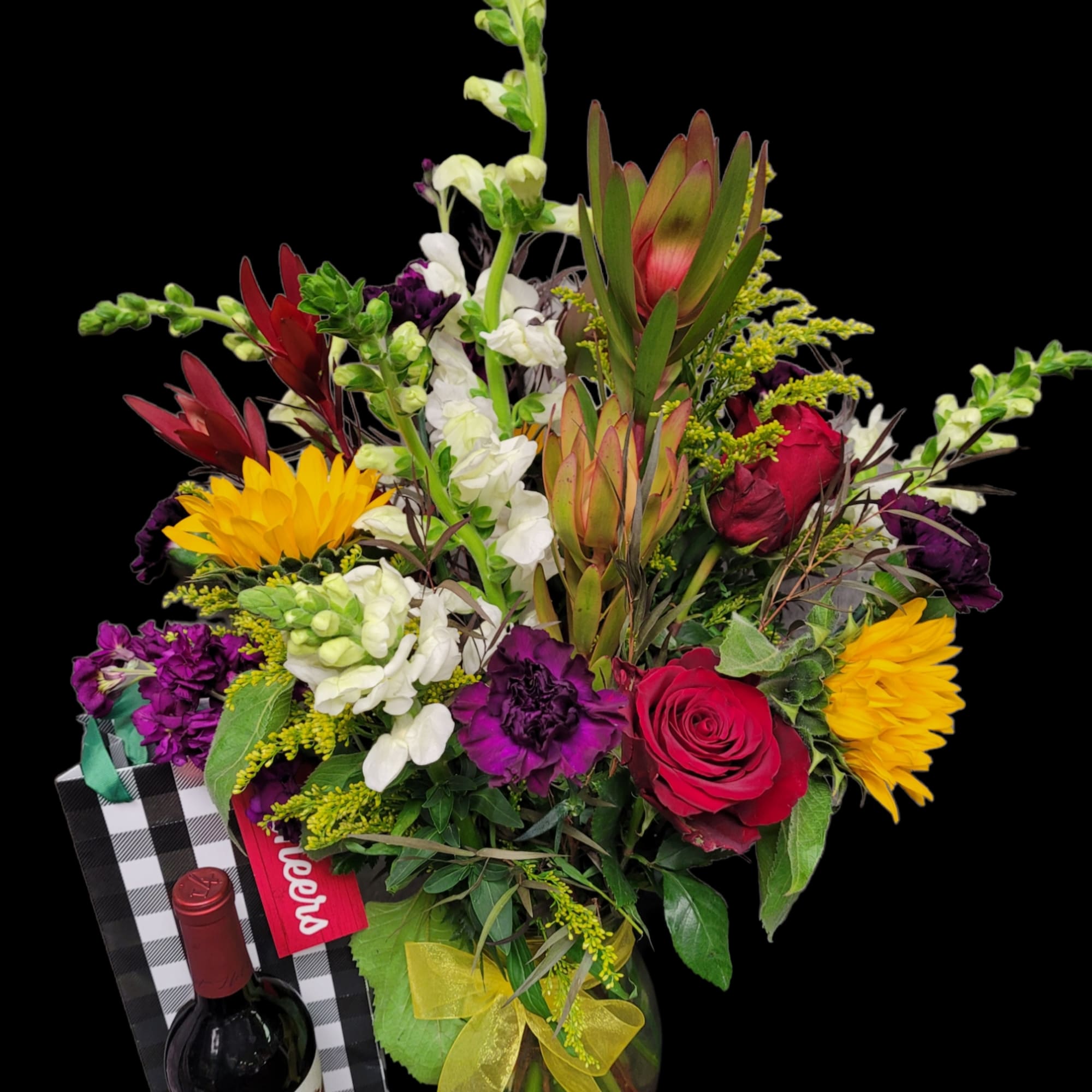 Mixed bouquet with roses, sunflowers, and snapdragons in a vase beside a wine bottle and gift bag