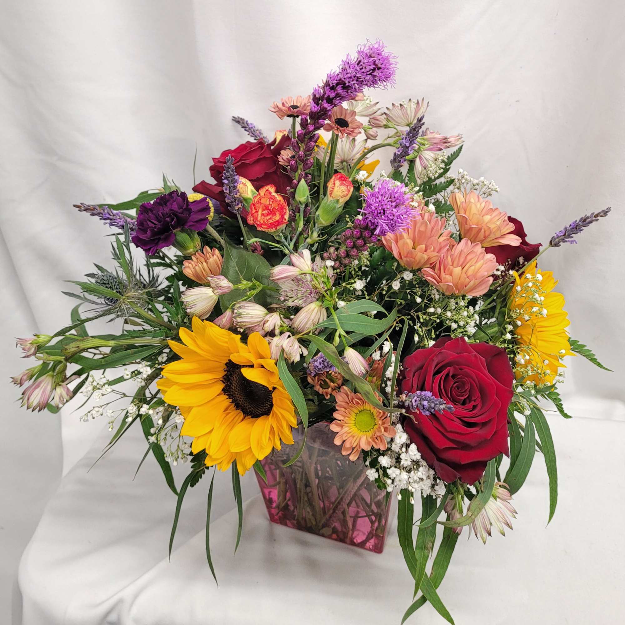 Mixed bouquet with sunflowers, red roses, and pink blooms in a pink glass vase