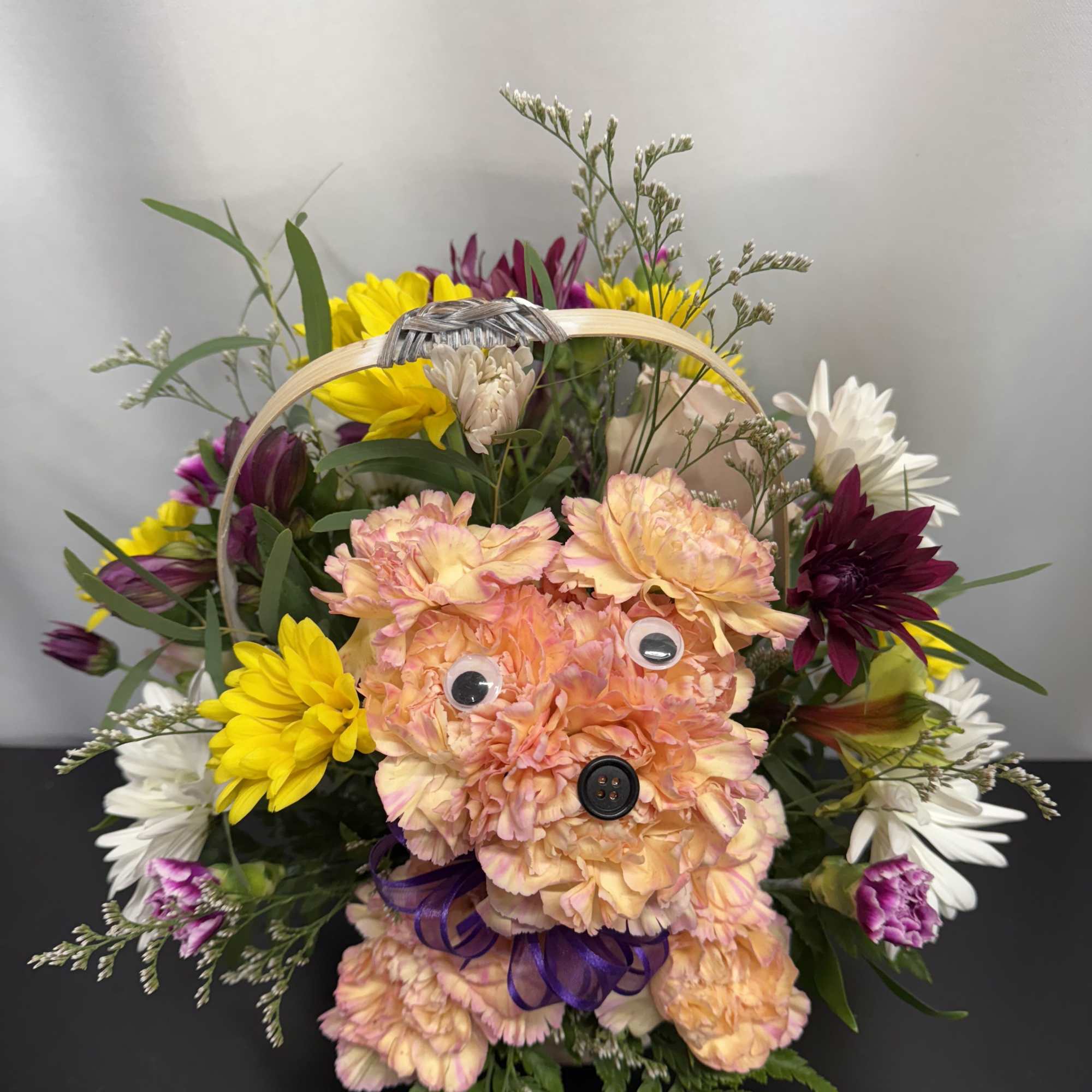Basket arrangement with peach carnation puppy figure surrounded by yellow, white, and purple mixed flowers