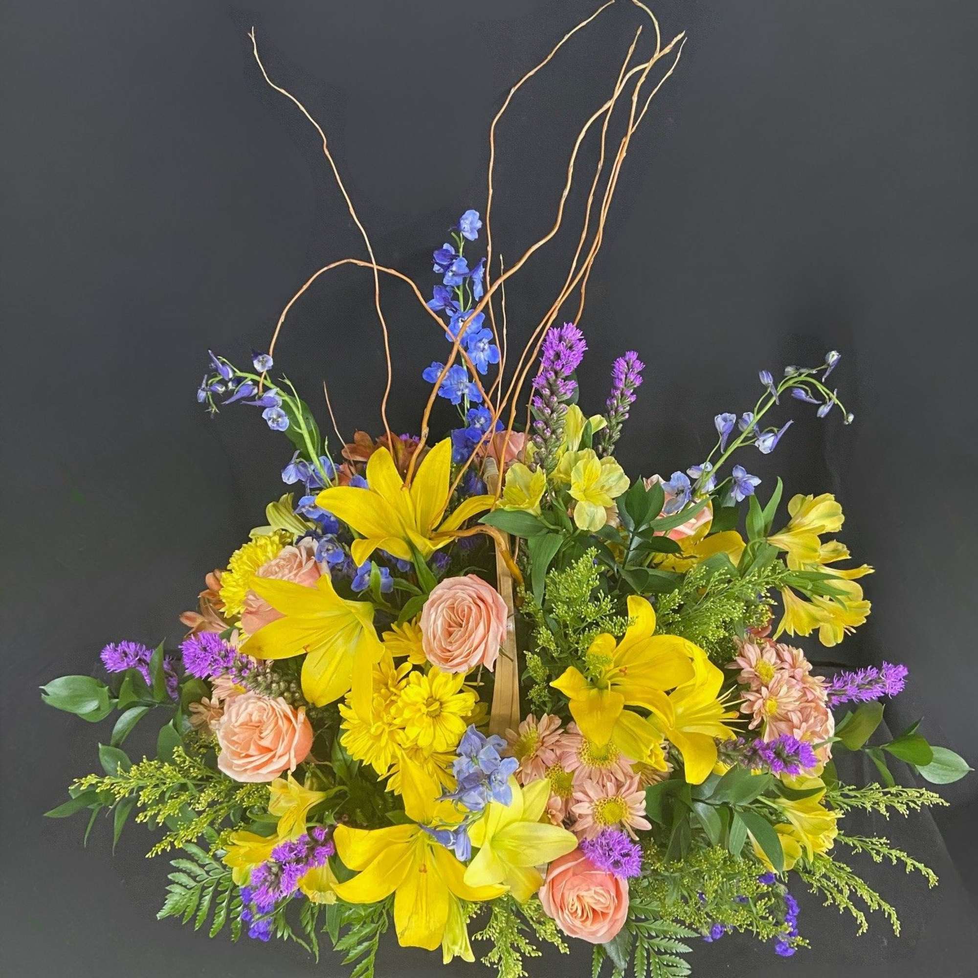 Basket arrangement of yellow lilies and mixed pastel flowers with curly branches on a dark background.