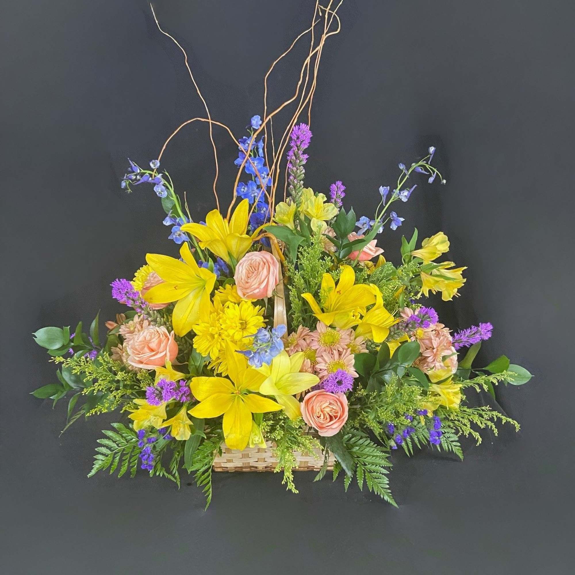 Bright basket arrangement with yellow lilies, peach roses, and mixed purple and blue flowers