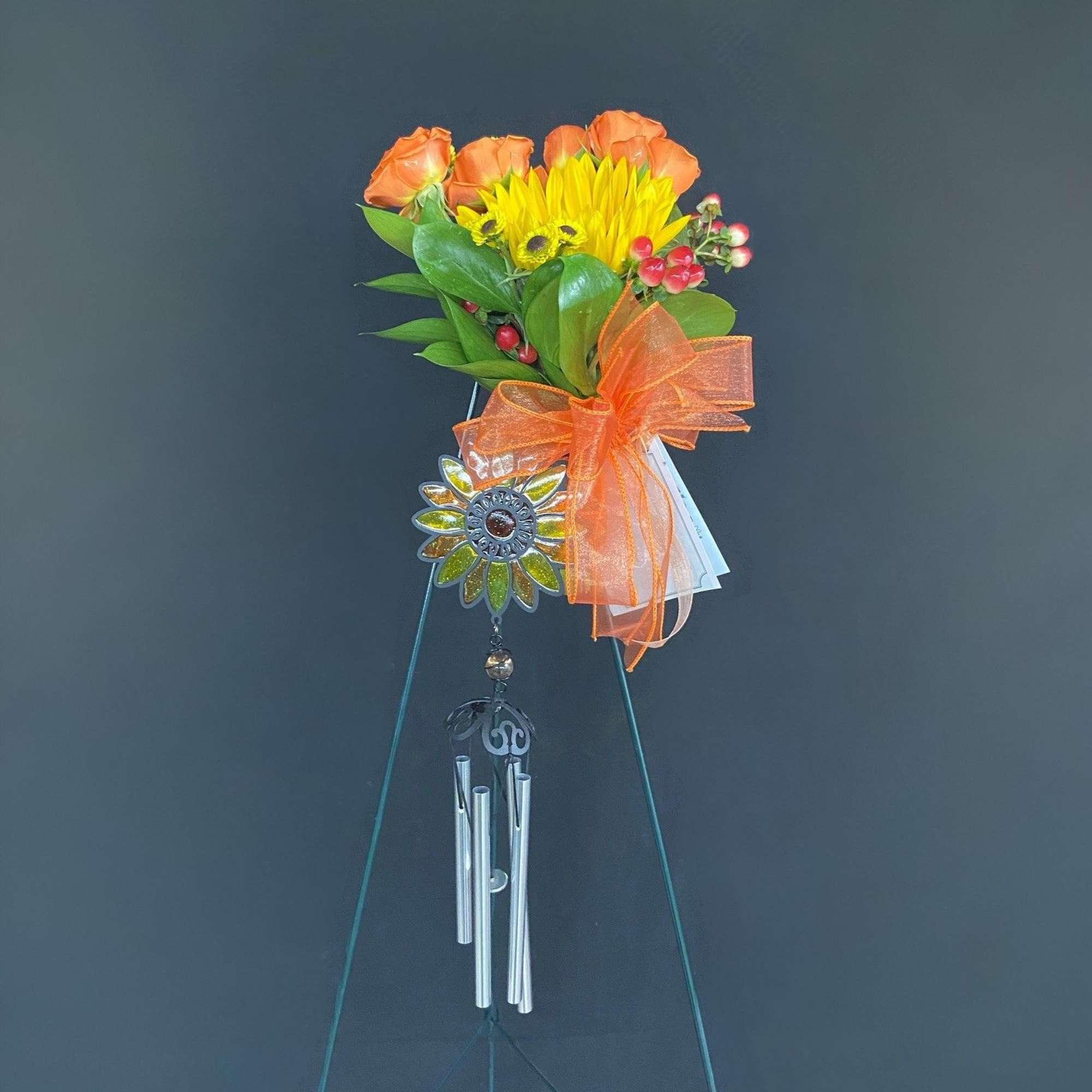 Orange roses and a yellow sunflower bouquet with an orange bow on a wind chime stand