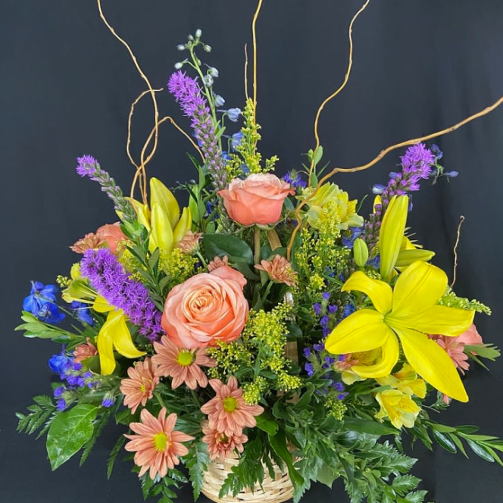 Basket arrangement with peach roses, yellow lilies, purple spikes, and blue flowers.
