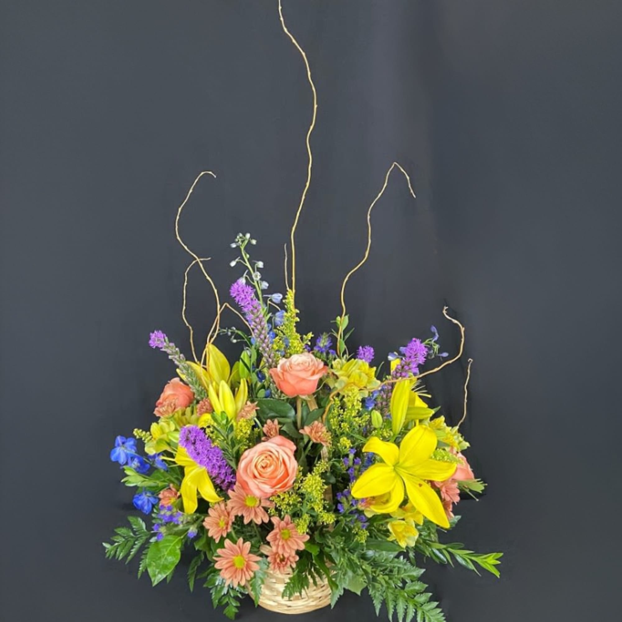 Tall mixed basket arrangement with yellow lilies, peach roses, and purple and blue accent flowers