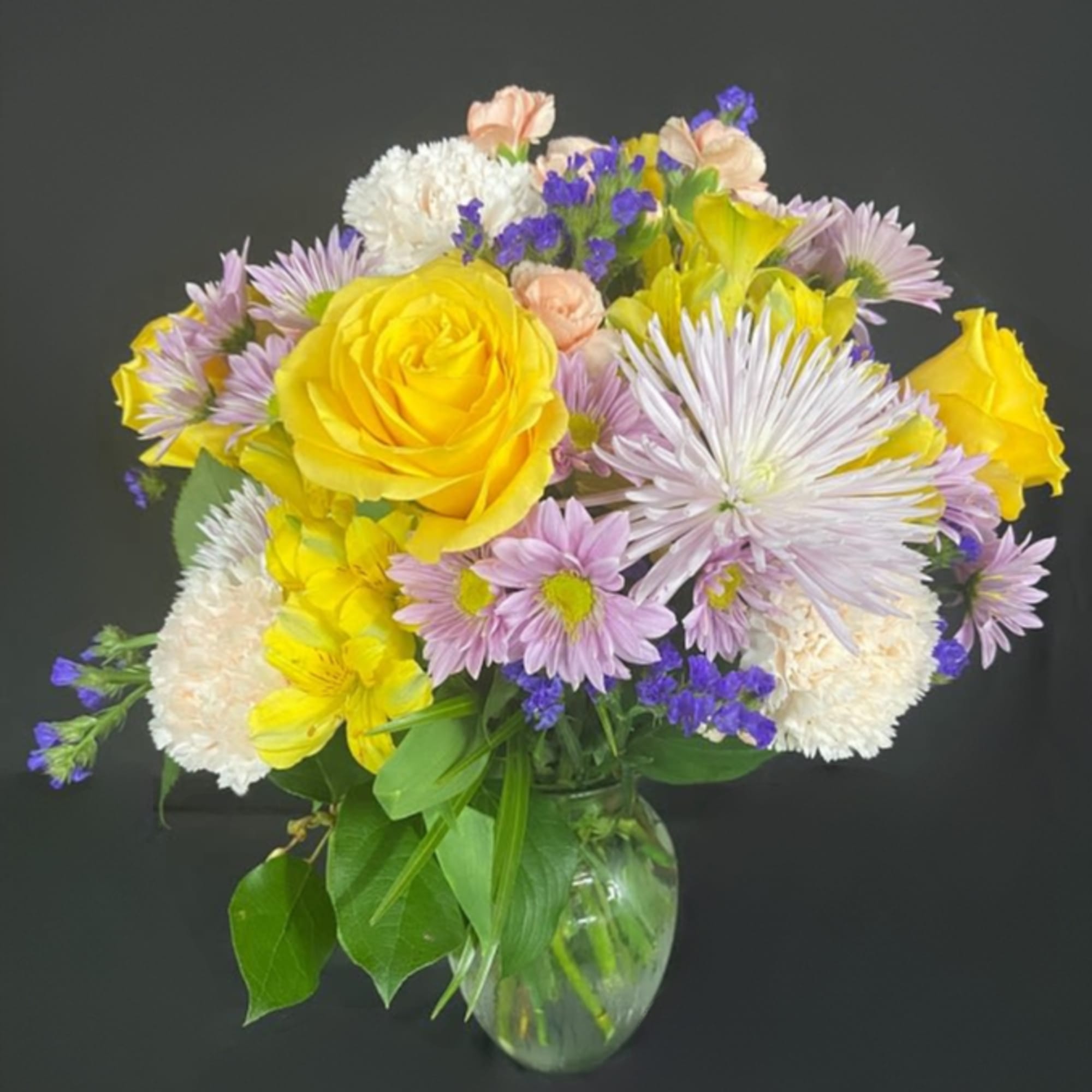 Mixed bouquet of yellow roses, lavender daisies, and white blooms in a clear glass vase
