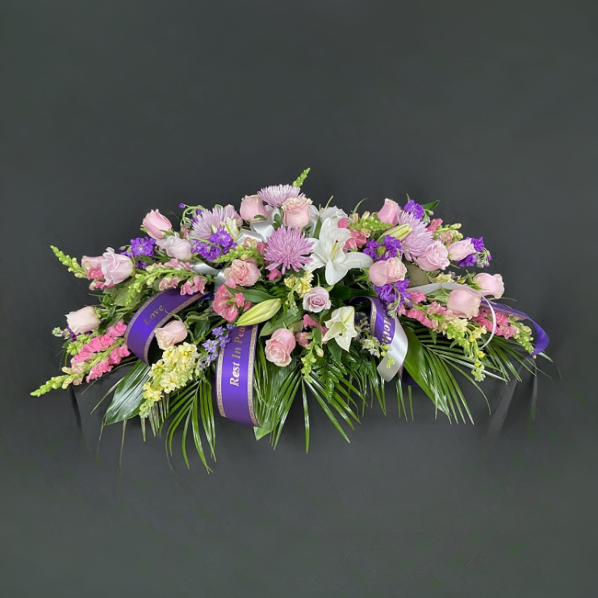 Pink and purple floral casket spray with white lilies and ribbon