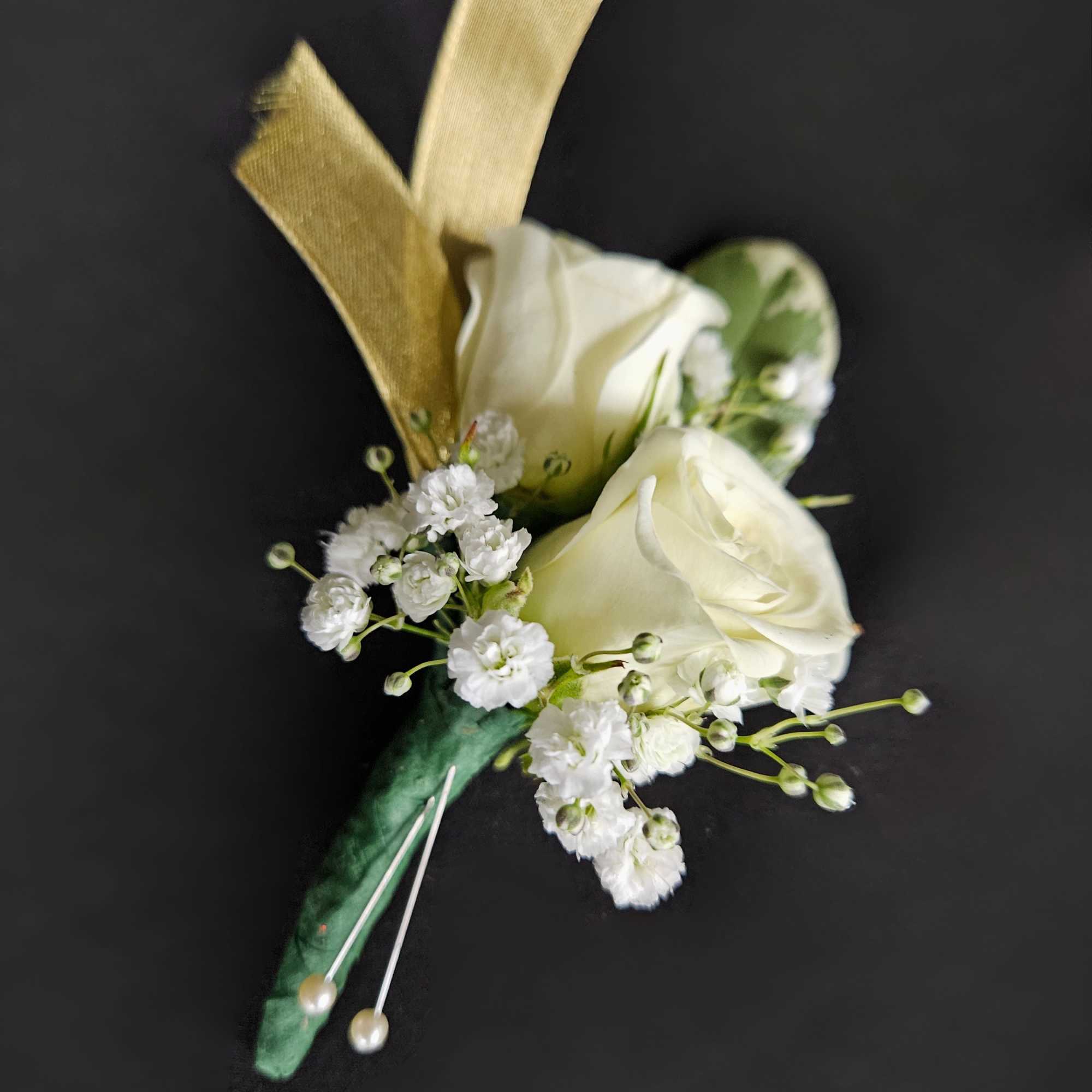 White rose boutonniere with tiny white filler flowers, gold ribbon, and pearl pins on a dark background.