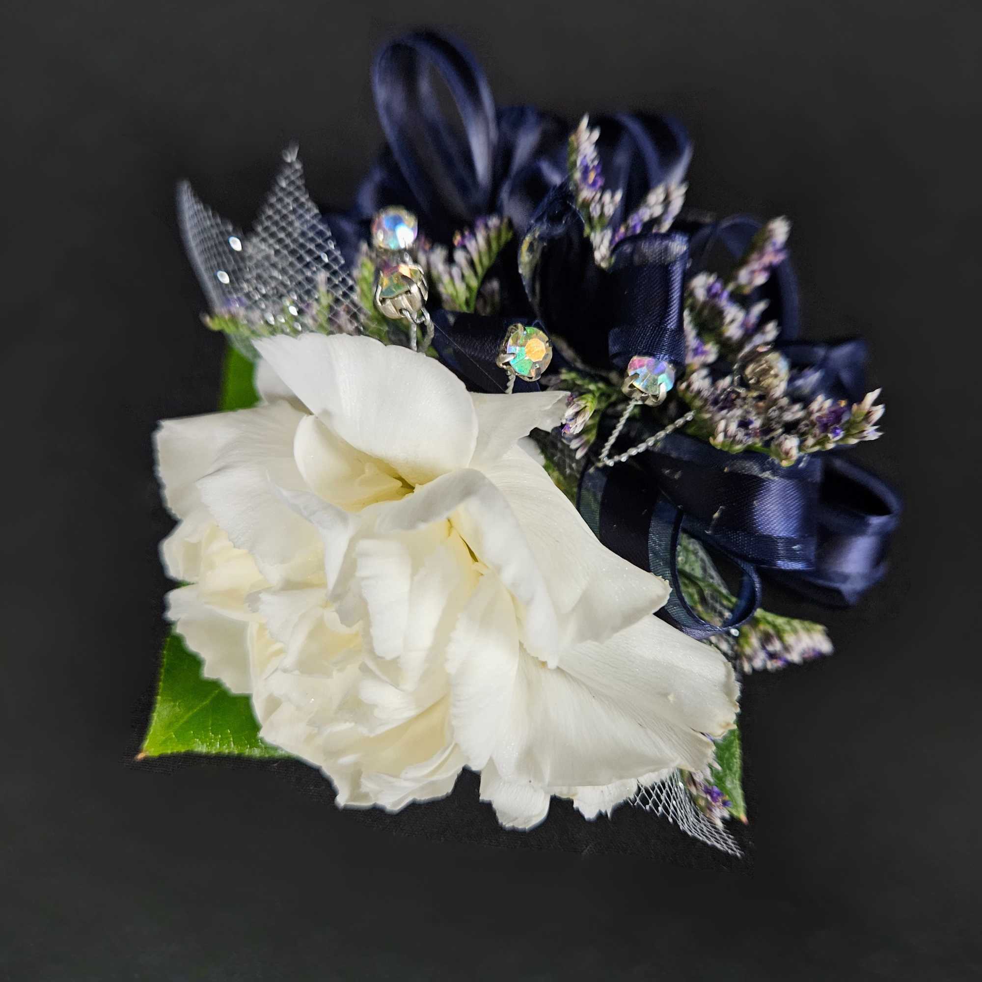 White carnation corsage with navy ribbon loops, tiny purple accents, and jeweled picks on a dark background.