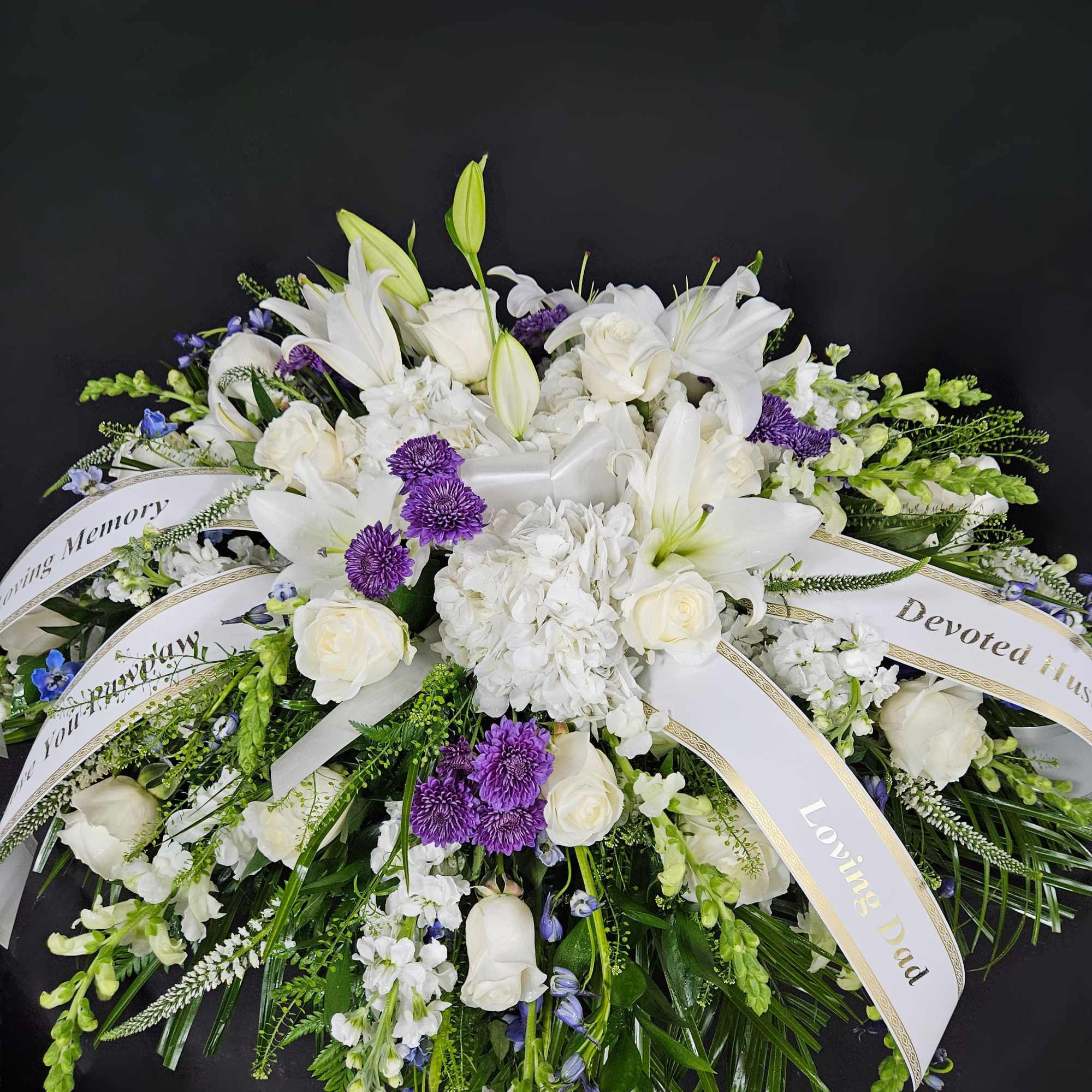 Large white and purple funeral spray with lilies, roses, and memorial ribbons on a dark background