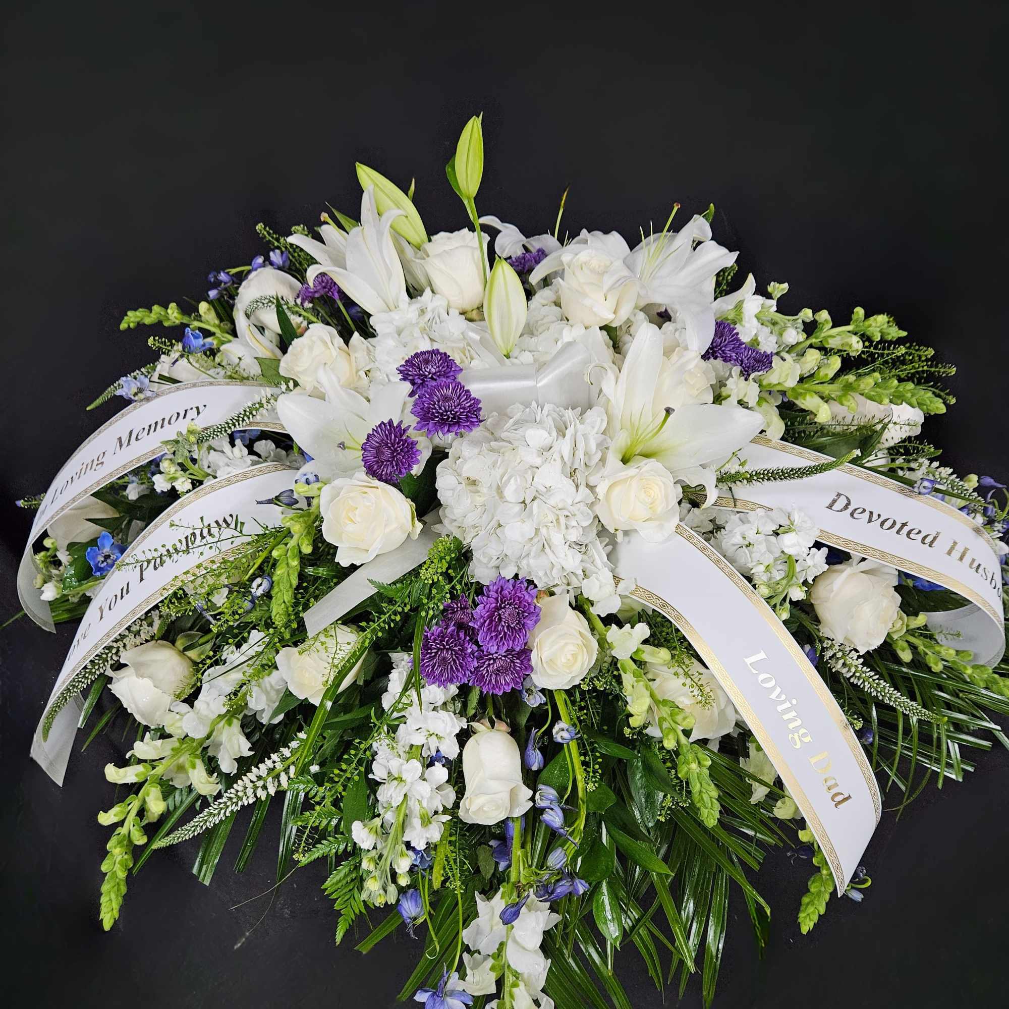 White and purple casket spray with lilies, roses, hydrangea, and memorial ribbons on a dark background.