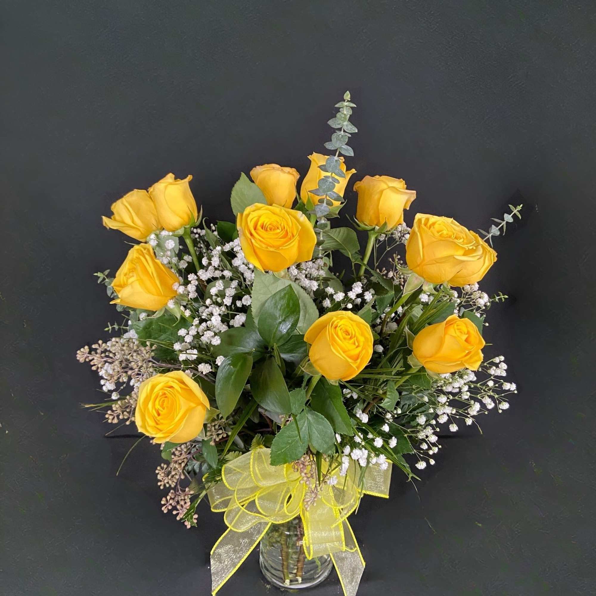 Arrangement of yellow roses with white filler flowers in a clear glass vase with a yellow bow.