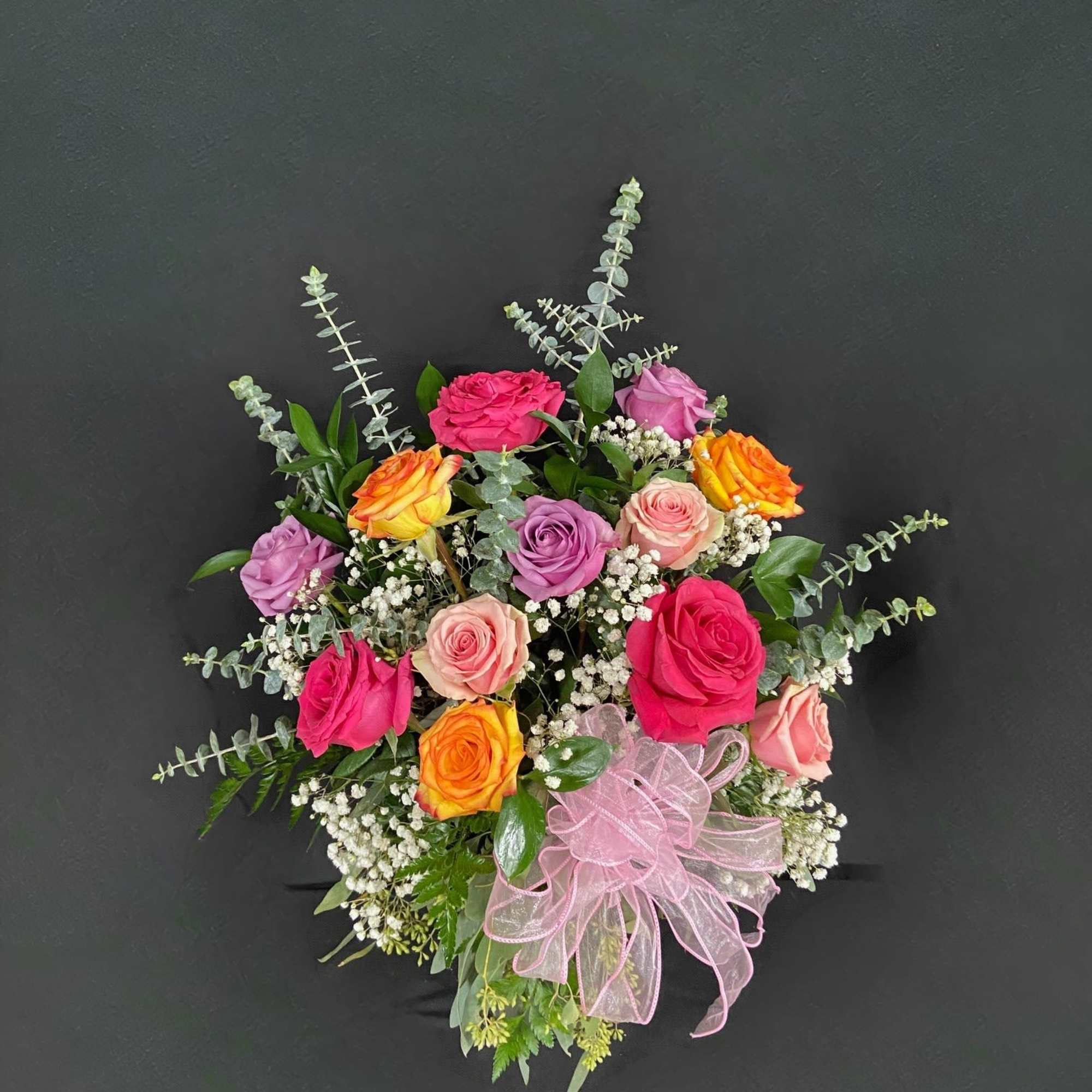 Mixed-color rose arrangement with baby’s breath and a pink ribbon bow on a dark background