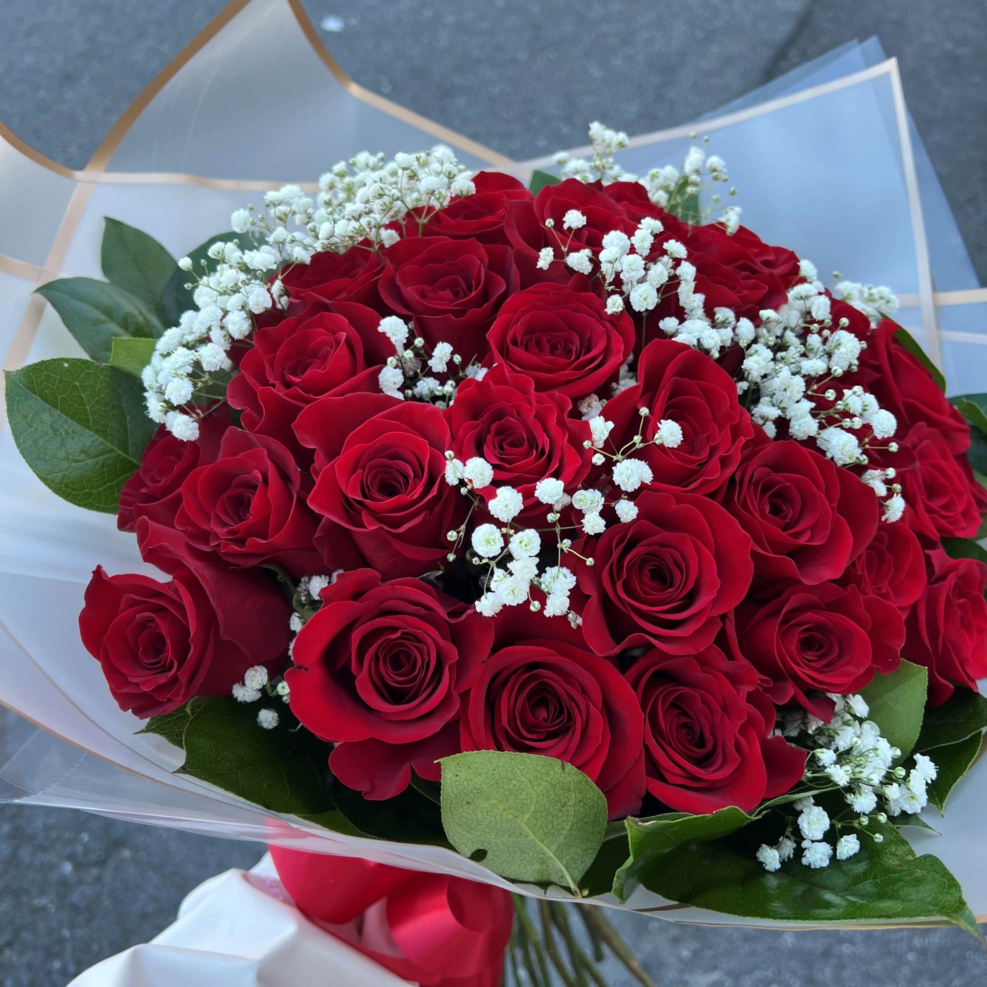 Handheld bouquet of red roses with small white filler flowers wrapped in sheer paper and tied with a red ribbon.