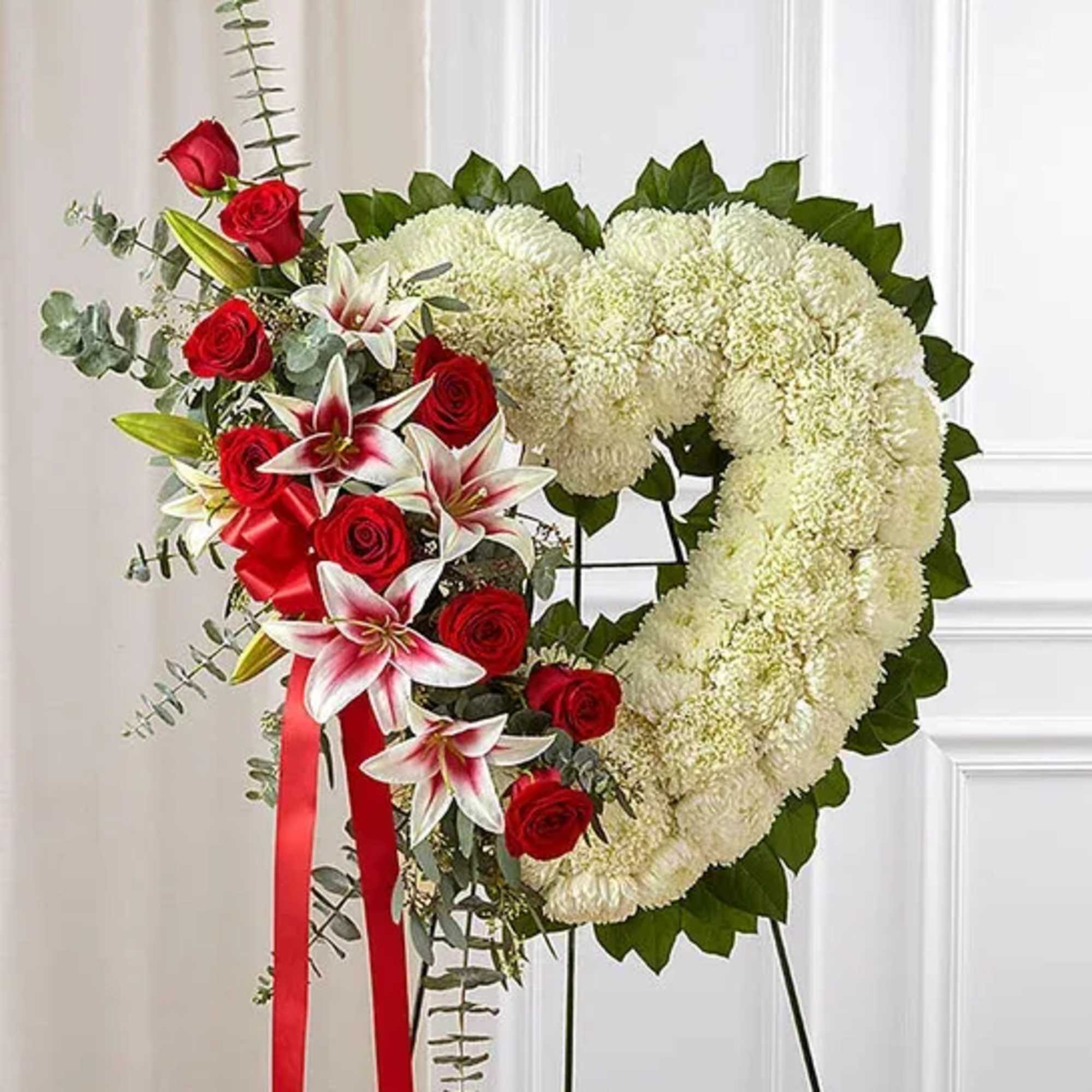 Heart-shaped white floral wreath with red roses, lilies, and ribbon on a standing easel.