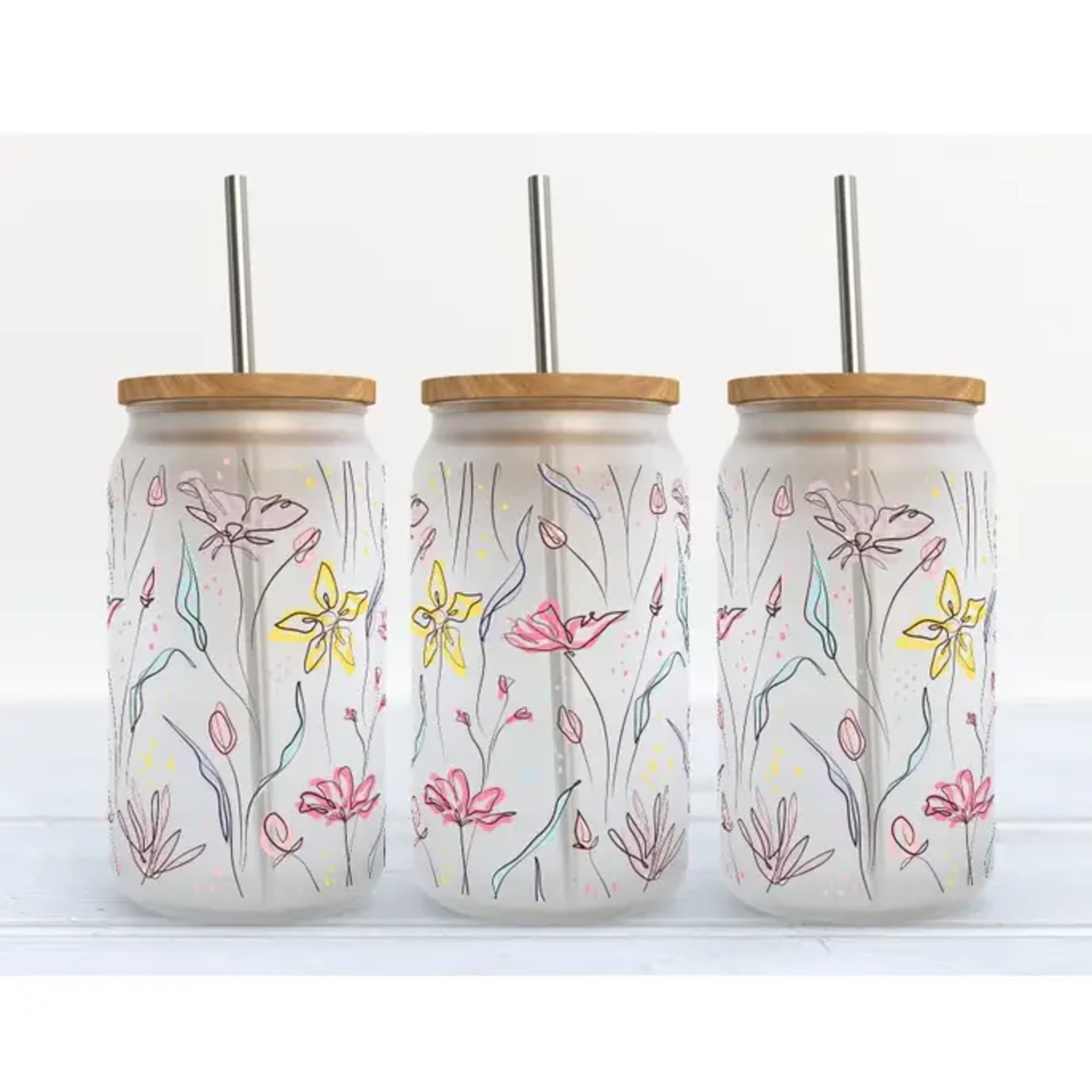 Three floral-patterned glass tumblers with metal straws and wooden lids