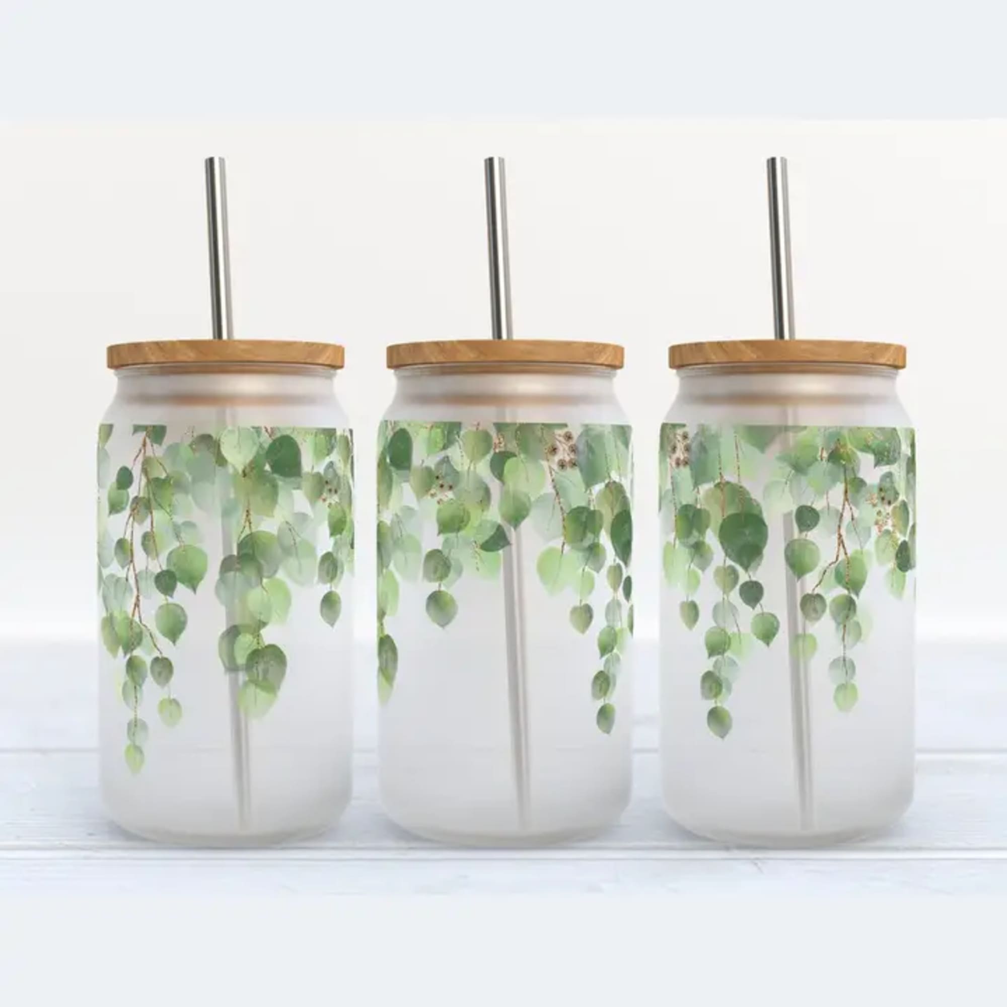 Three clear tumblers with wood lids and metal straws, printed with hanging green leaves.