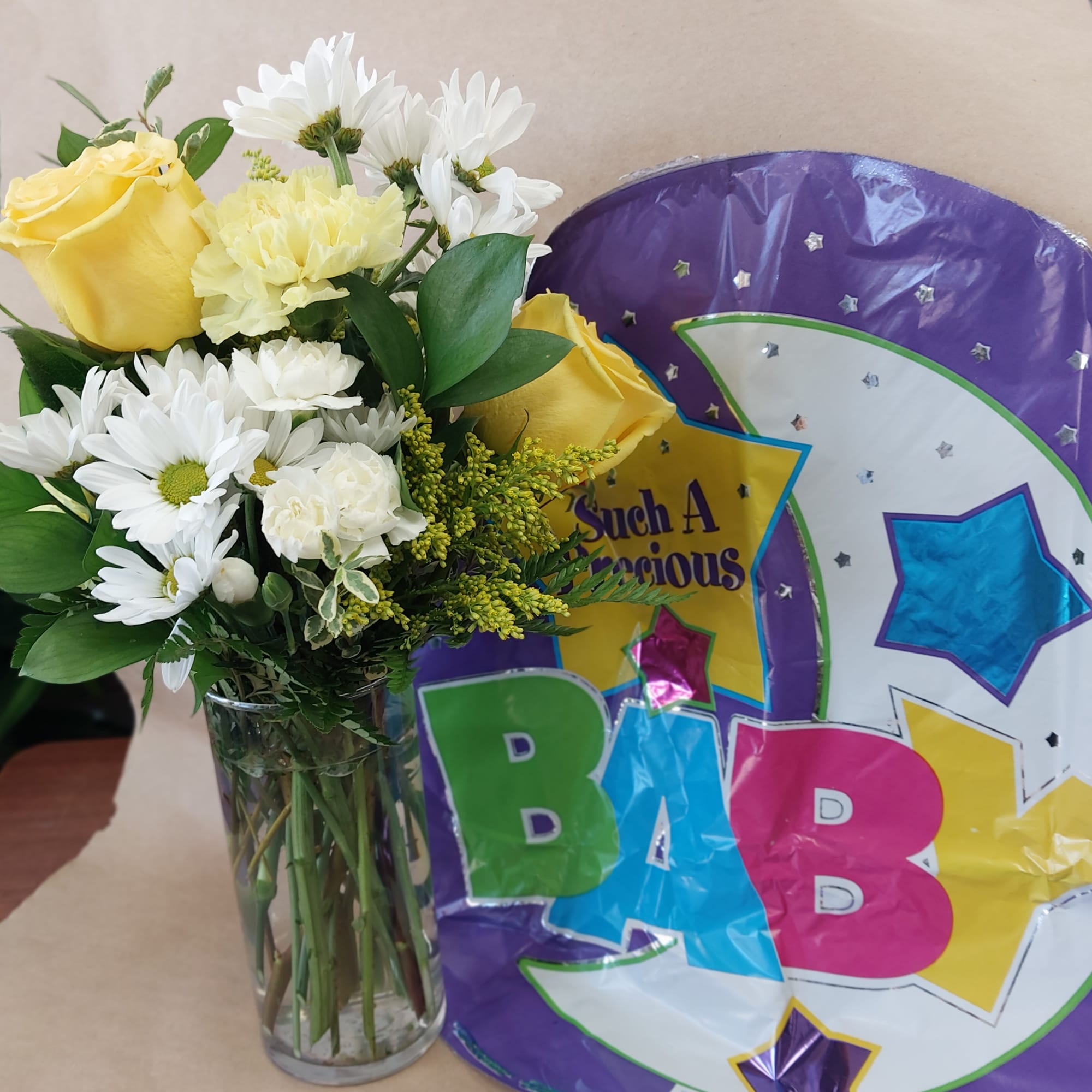 Yellow roses and white daisies in a glass vase beside a purple baby balloon