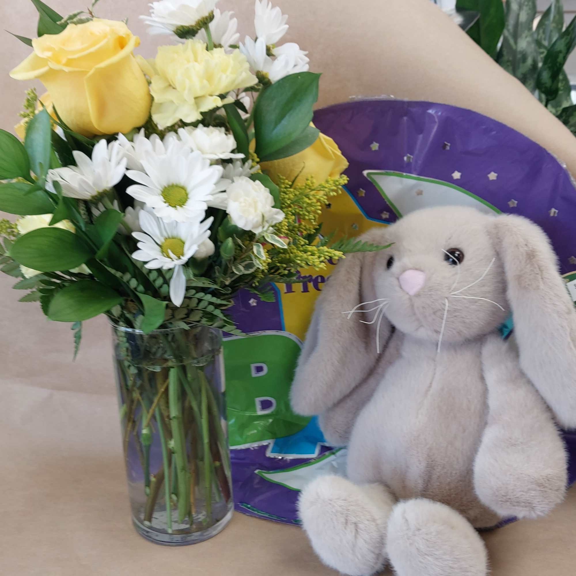 Yellow roses and white daisies in a glass vase beside a plush bunny