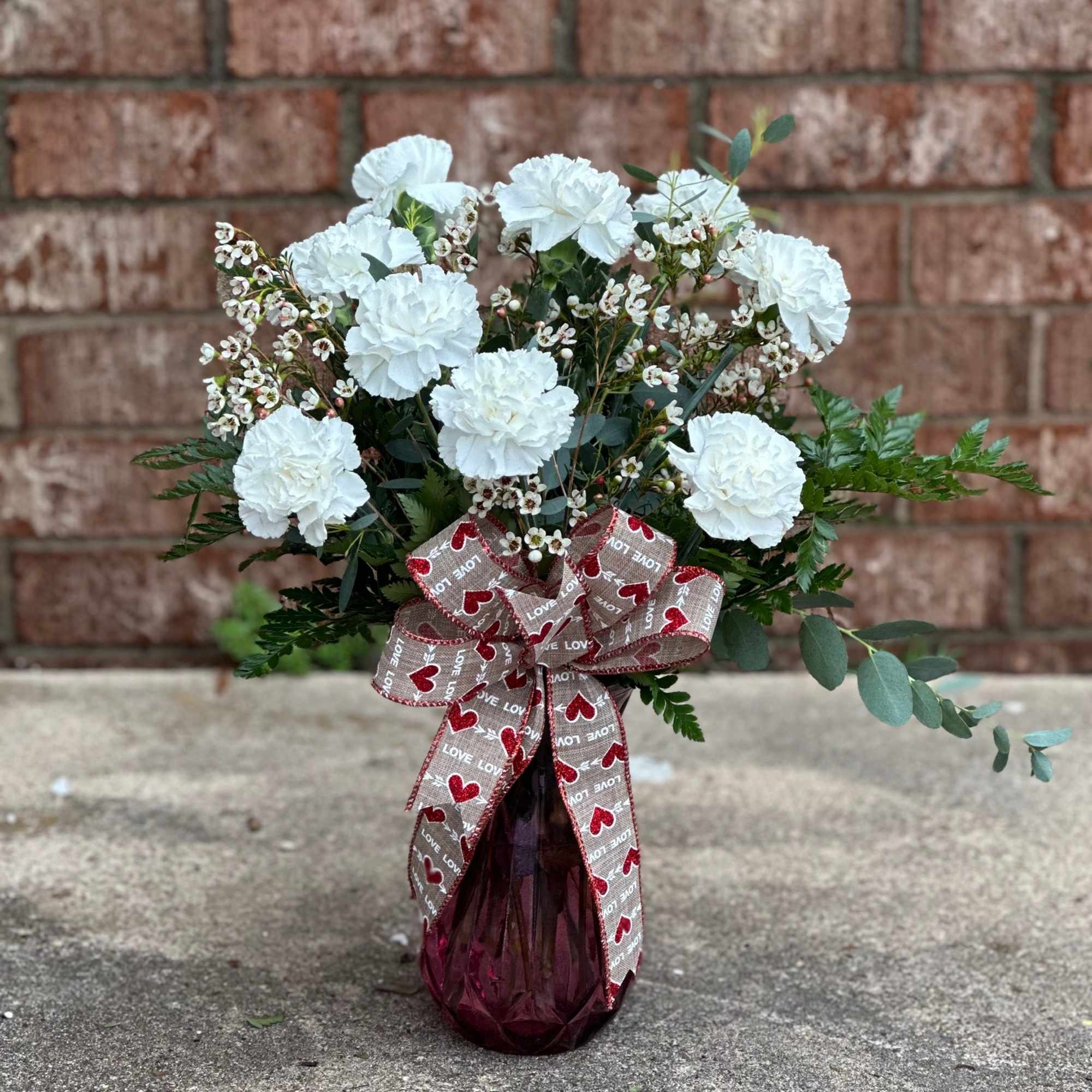 White carnations in a burgundy vase with a heart-patterned ribbon