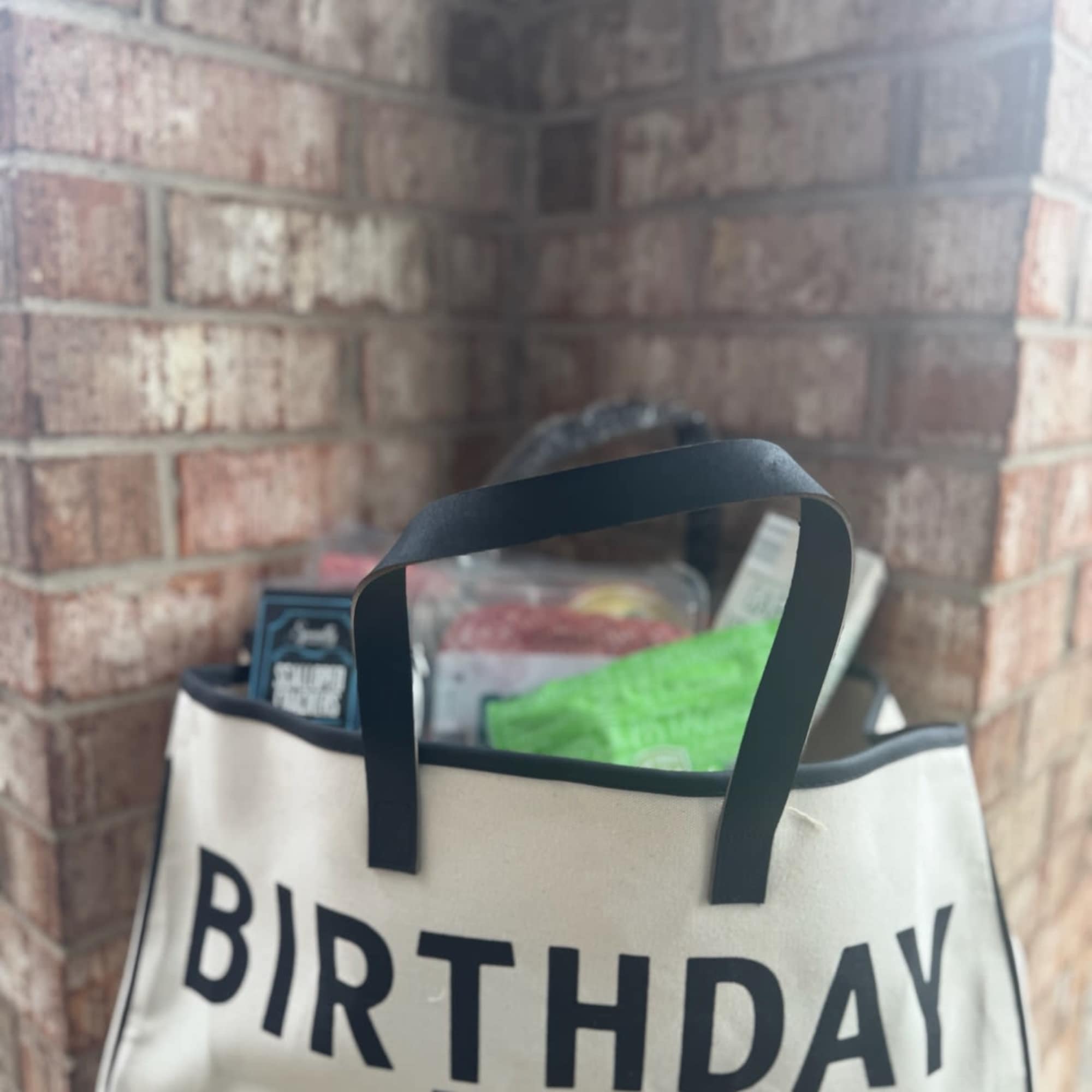 A birthday tote bag filled with gift items sits against a brick wall.