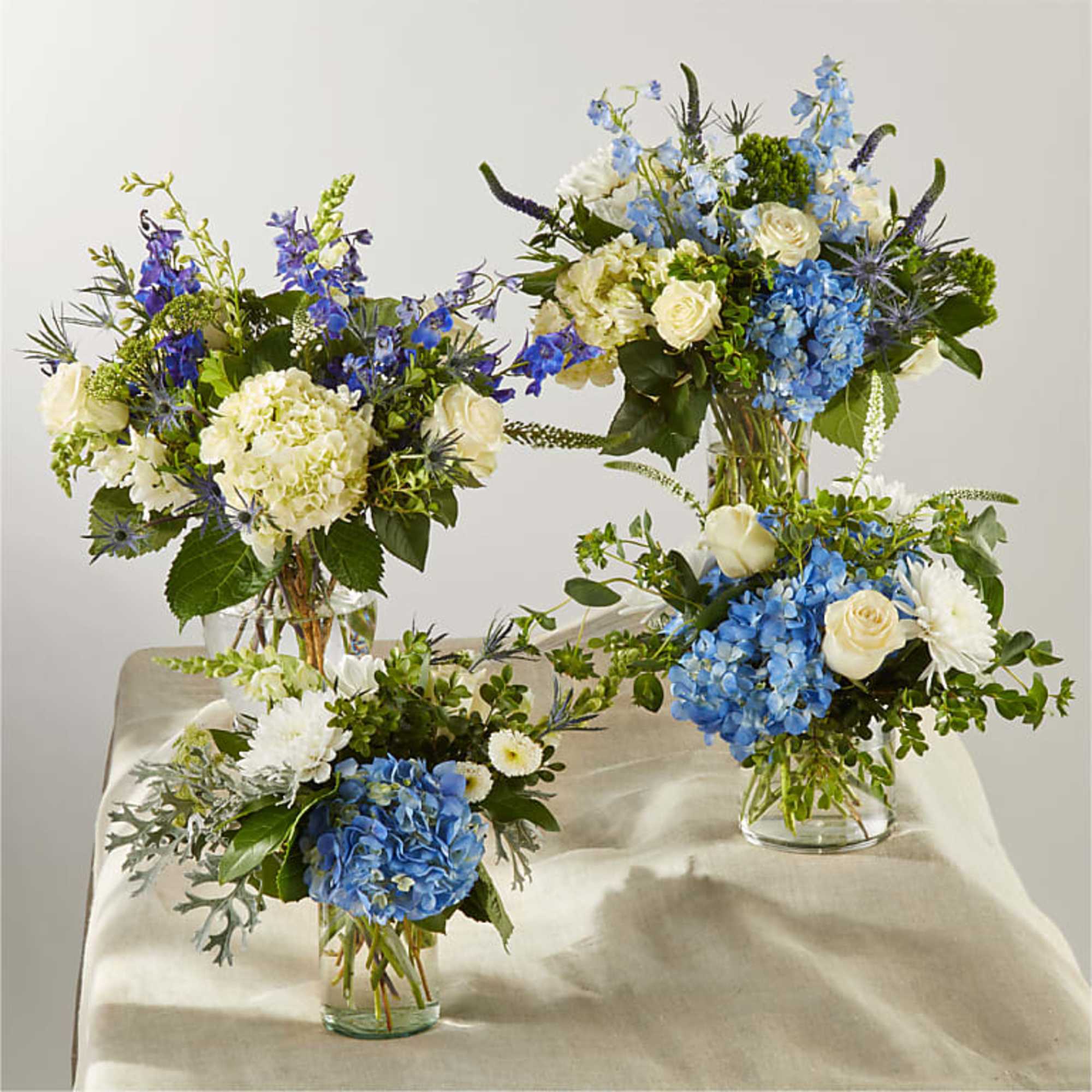 Four blue and white floral arrangements in clear glass vases on a draped table