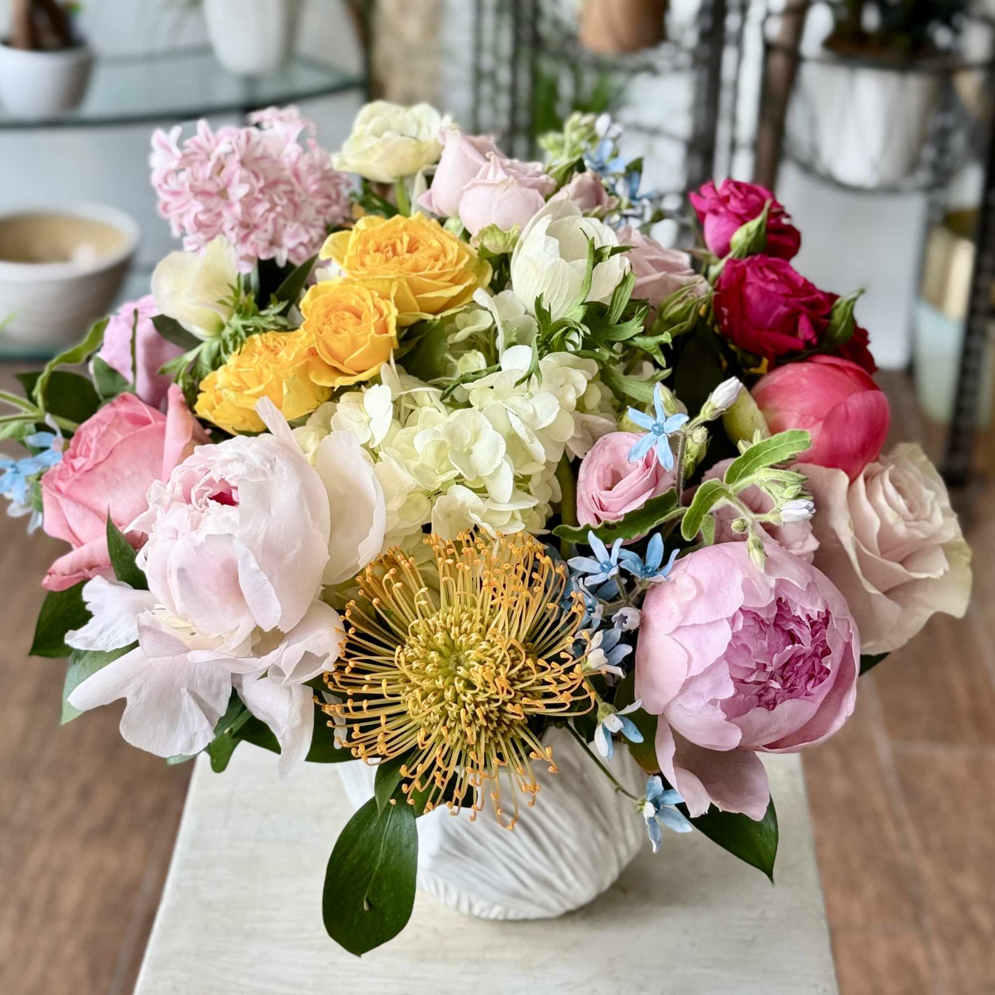 Low pastel arrangement of mixed pink peonies, roses, hydrangeas, and pincushion protea in a white ceramic vase