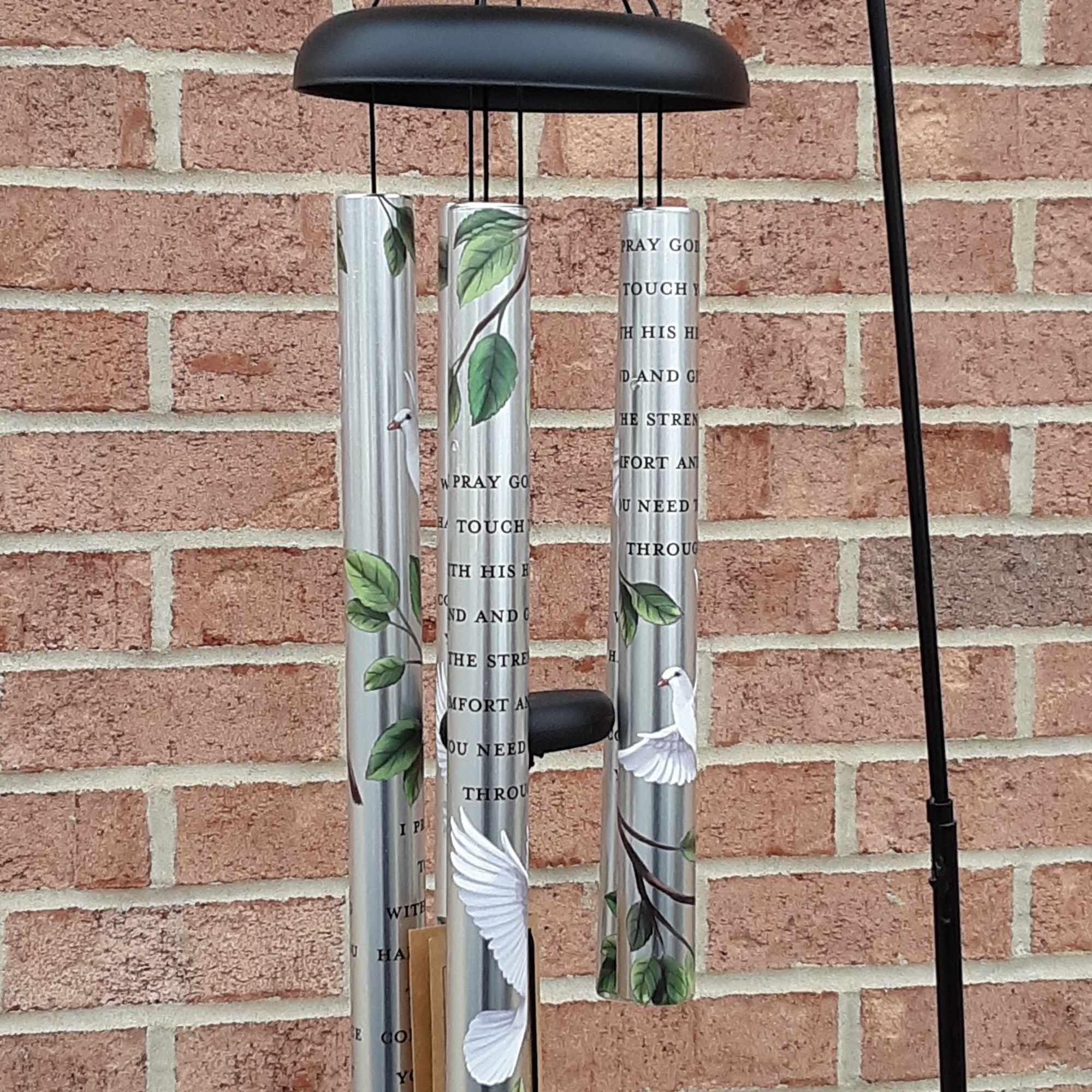 Black metal wind chime with silver tubes and bird designs hanging on a brick wall.