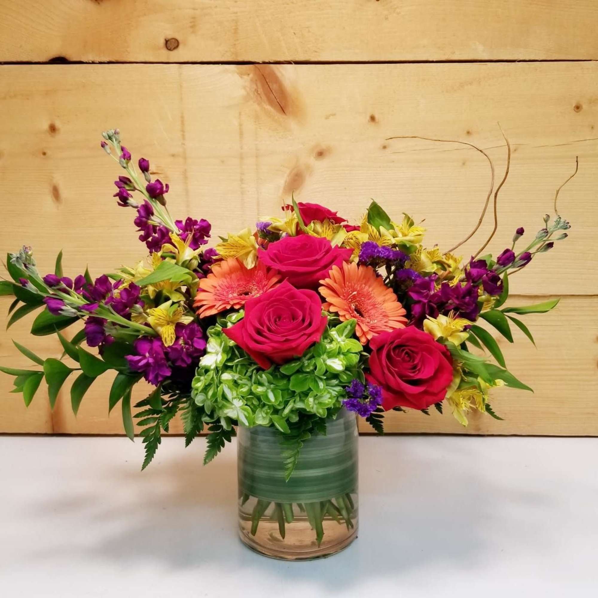 Mixed bouquet of pink roses, orange gerberas, and purple flowers in a glass vase