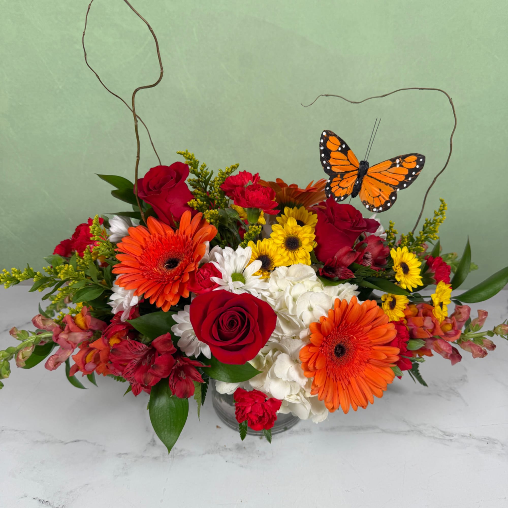 Mixed bouquet with red roses, orange gerberas, white hydrangeas, and a butterfly pick in a glass vase