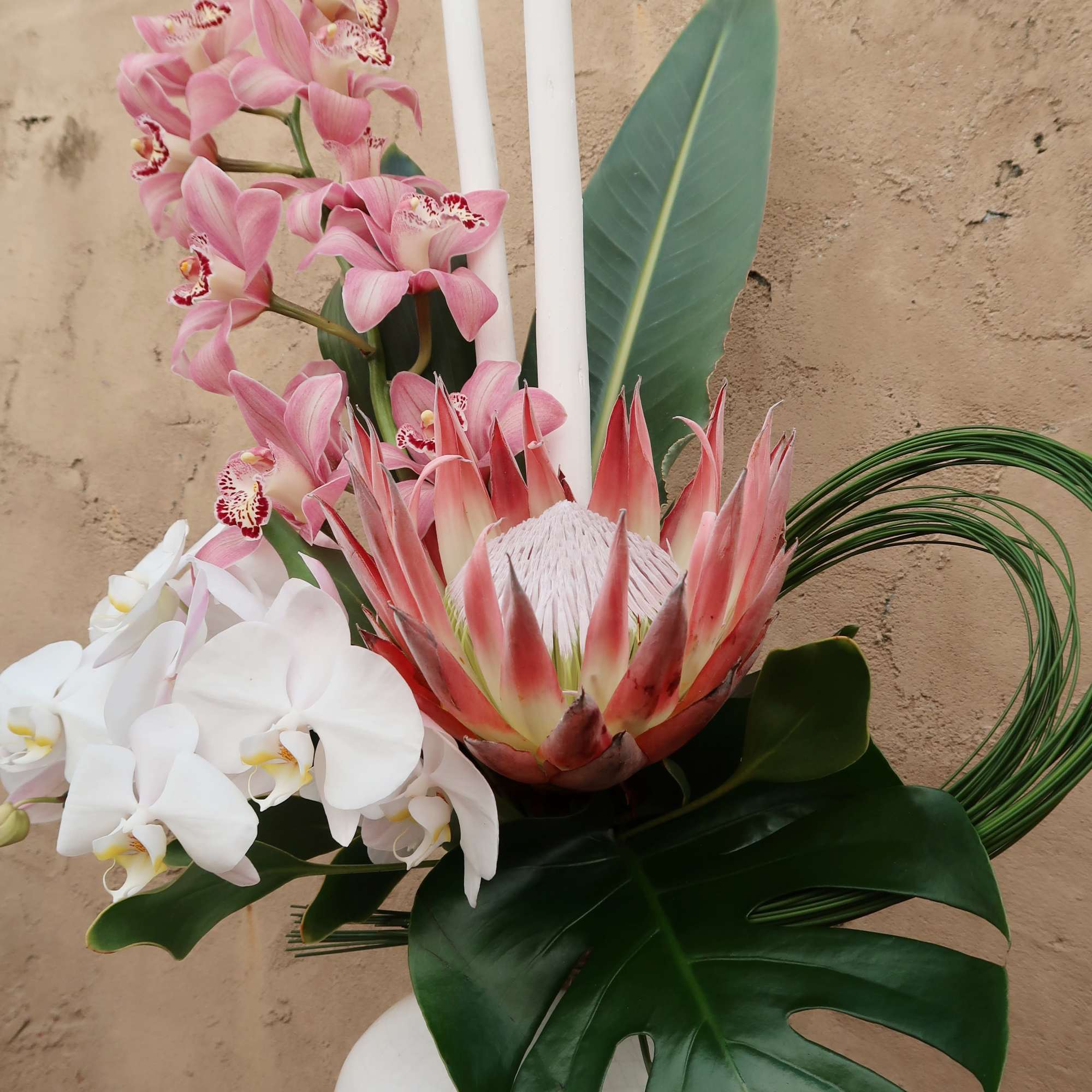 Pink orchids and a protea arranged with white candles in a vase.