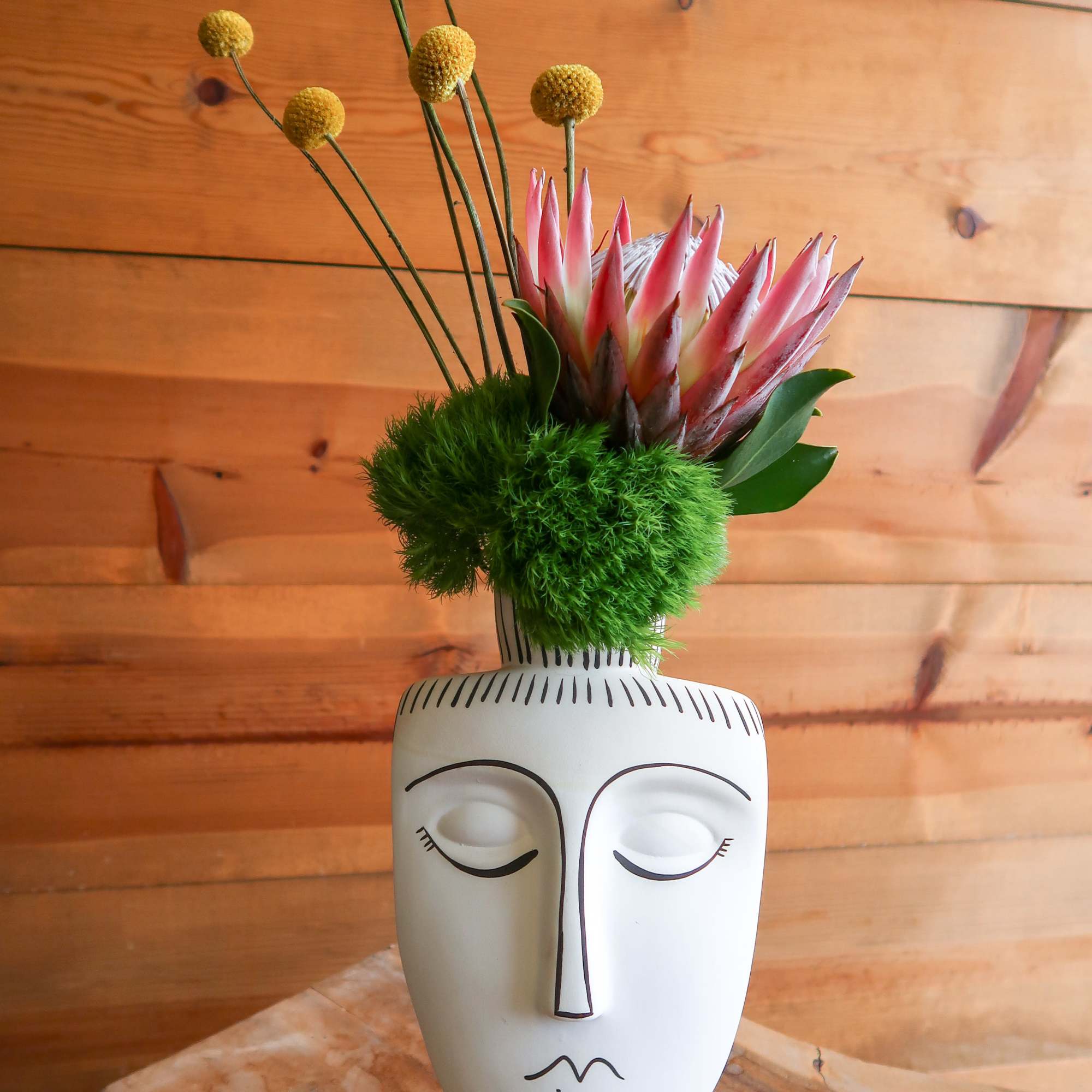 Pink protea and yellow pom-pom stems in a white face vase