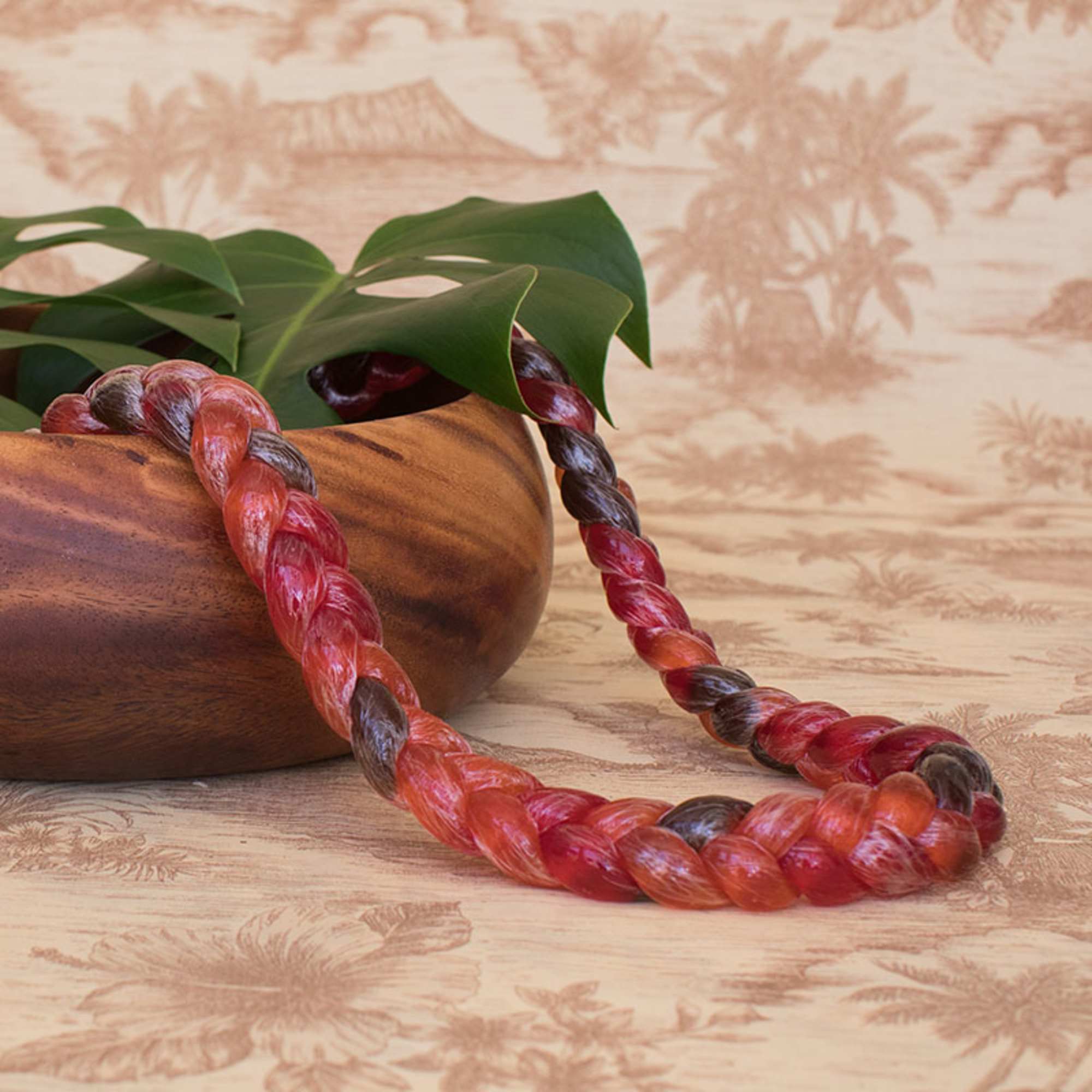 Braided red and orange lei draped over a wooden bowl with large tropical leaves.