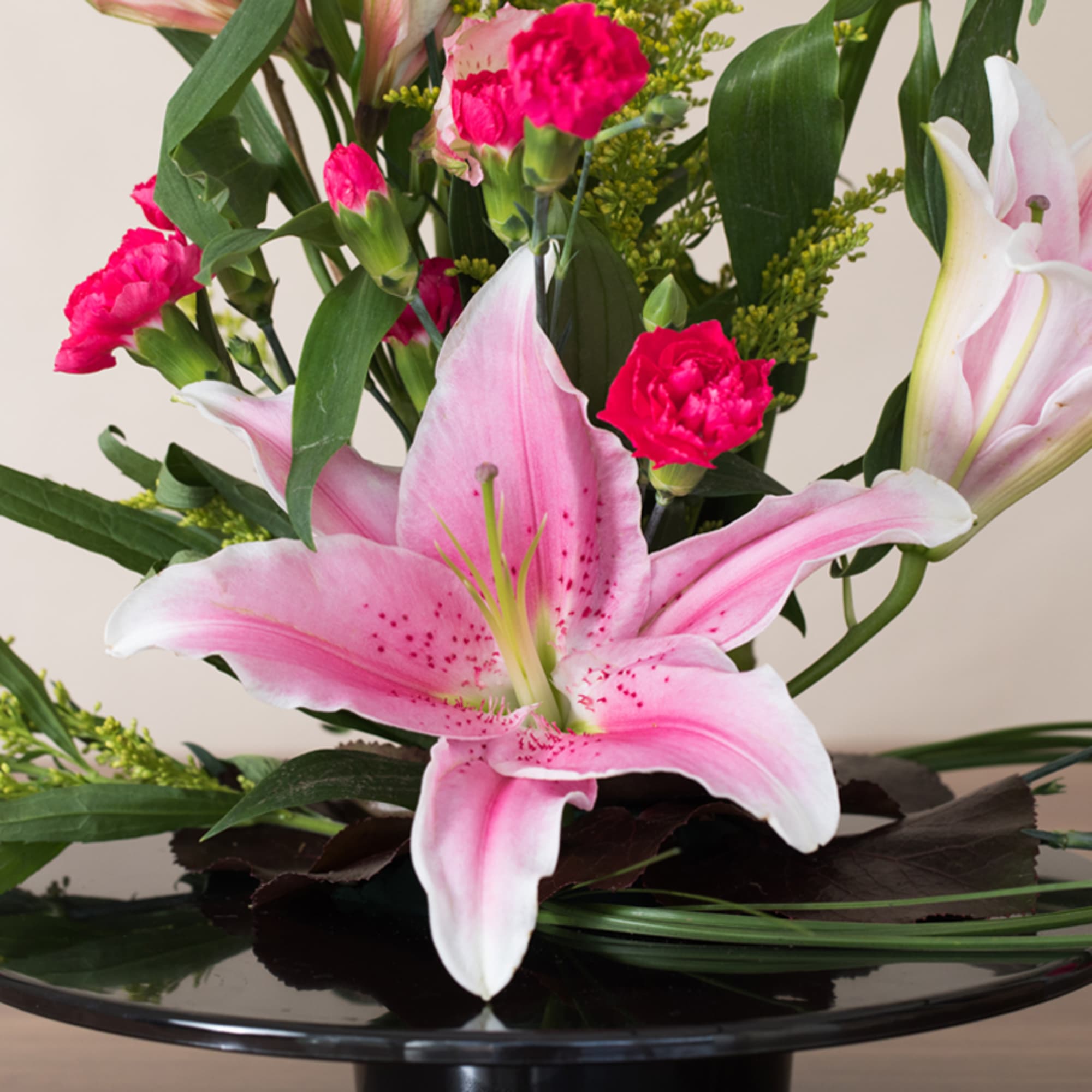 Pink lilies and bright pink carnations arranged in a low black pedestal dish.