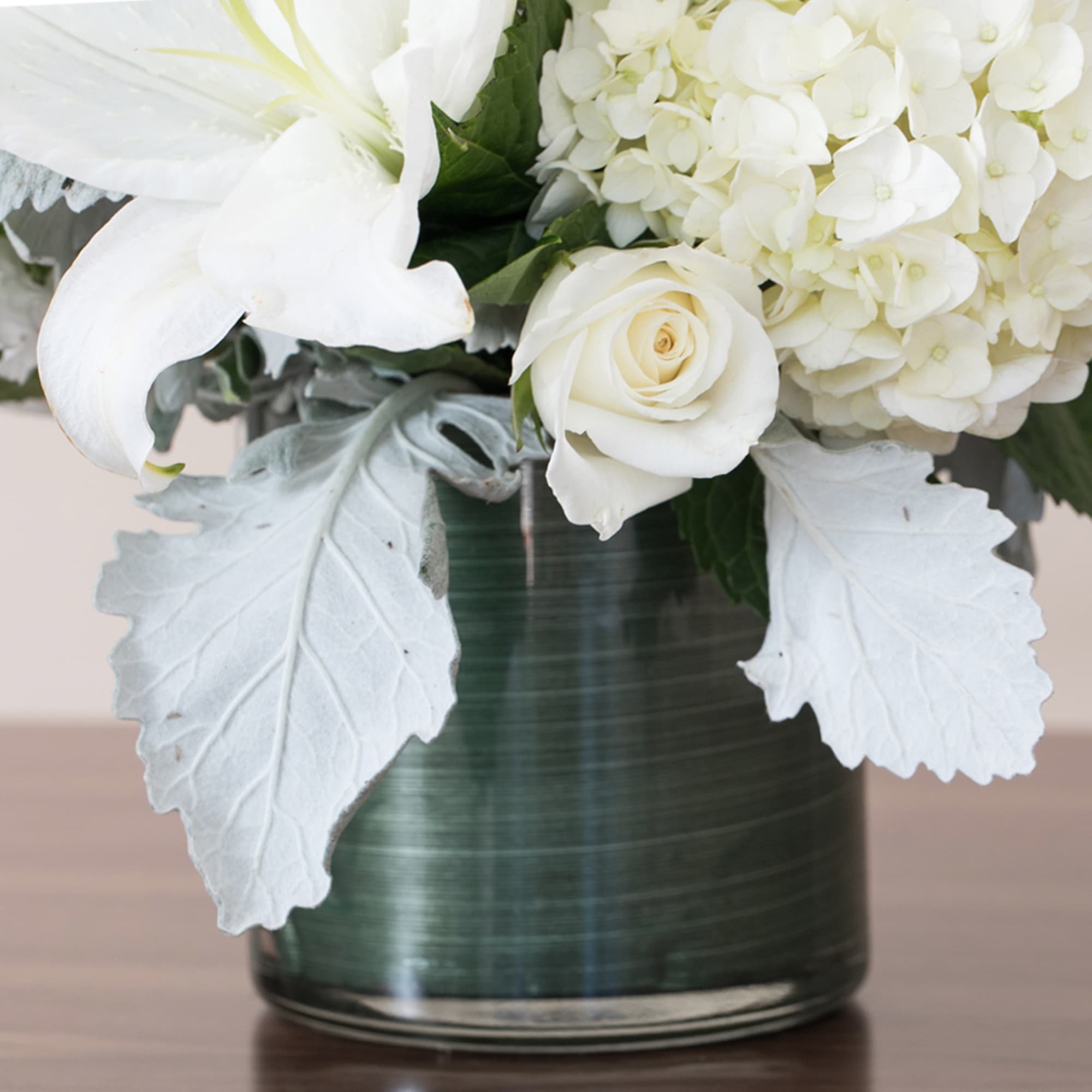 White lilies, roses, and hydrangea blooms in a dark green glass cylinder vase with soft silver foliage.