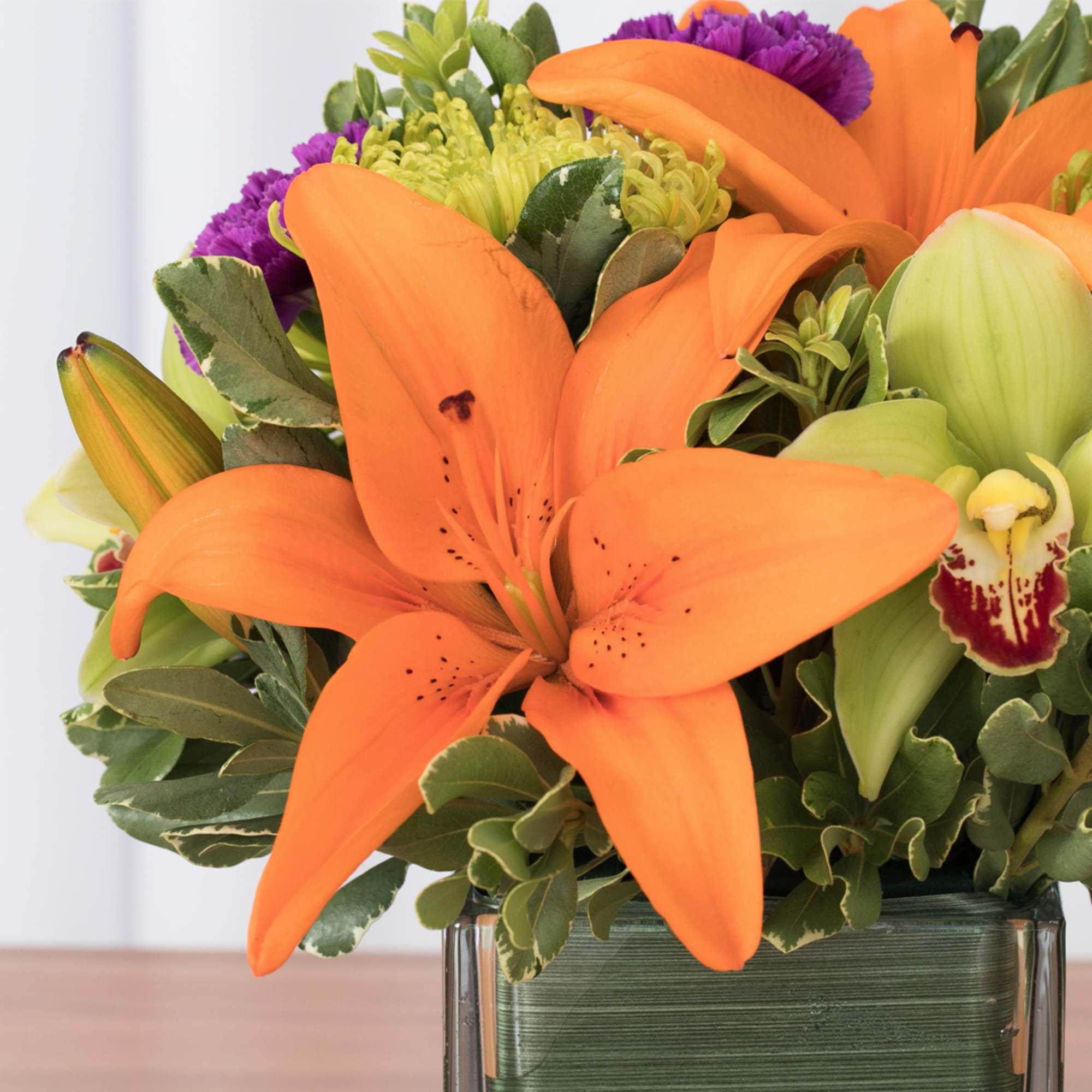 Bright orange lilies with green orchids and purple accents in a square glass vase