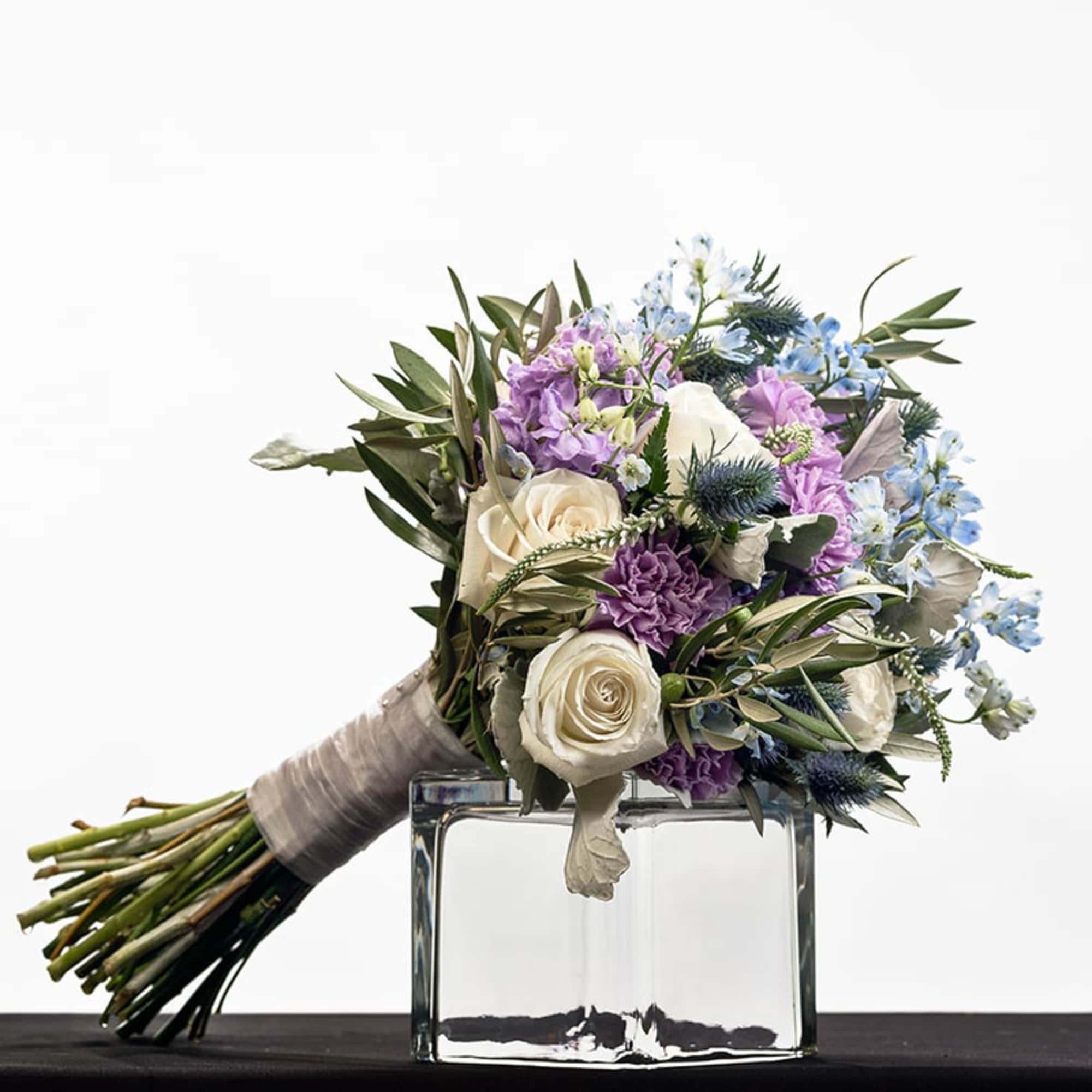 Hand-tied bouquet of white roses with lavender and blue flowers resting on a clear glass cube vase