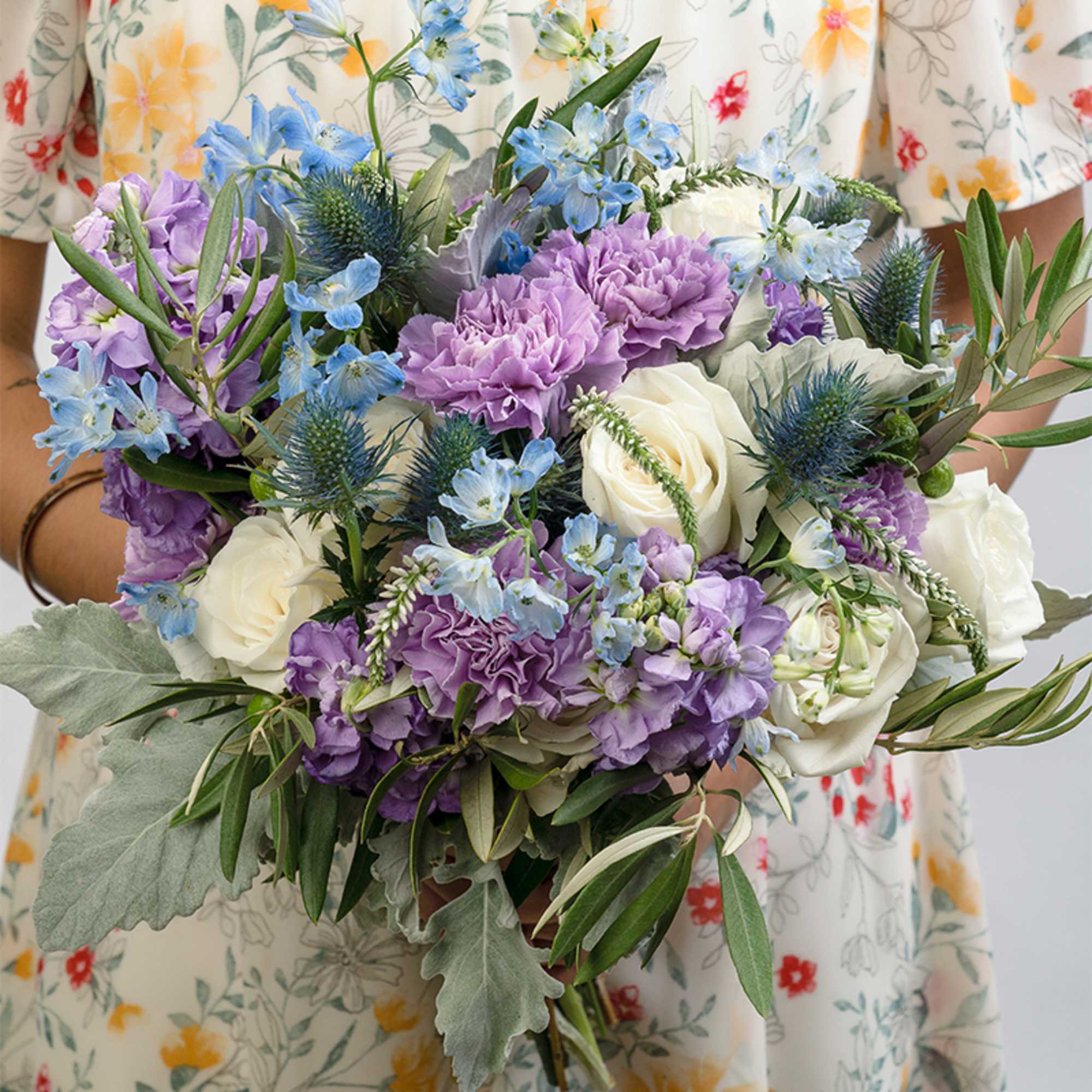 Hand-tied bouquet of white roses, lavender blooms, and blue flowers held against a floral dress