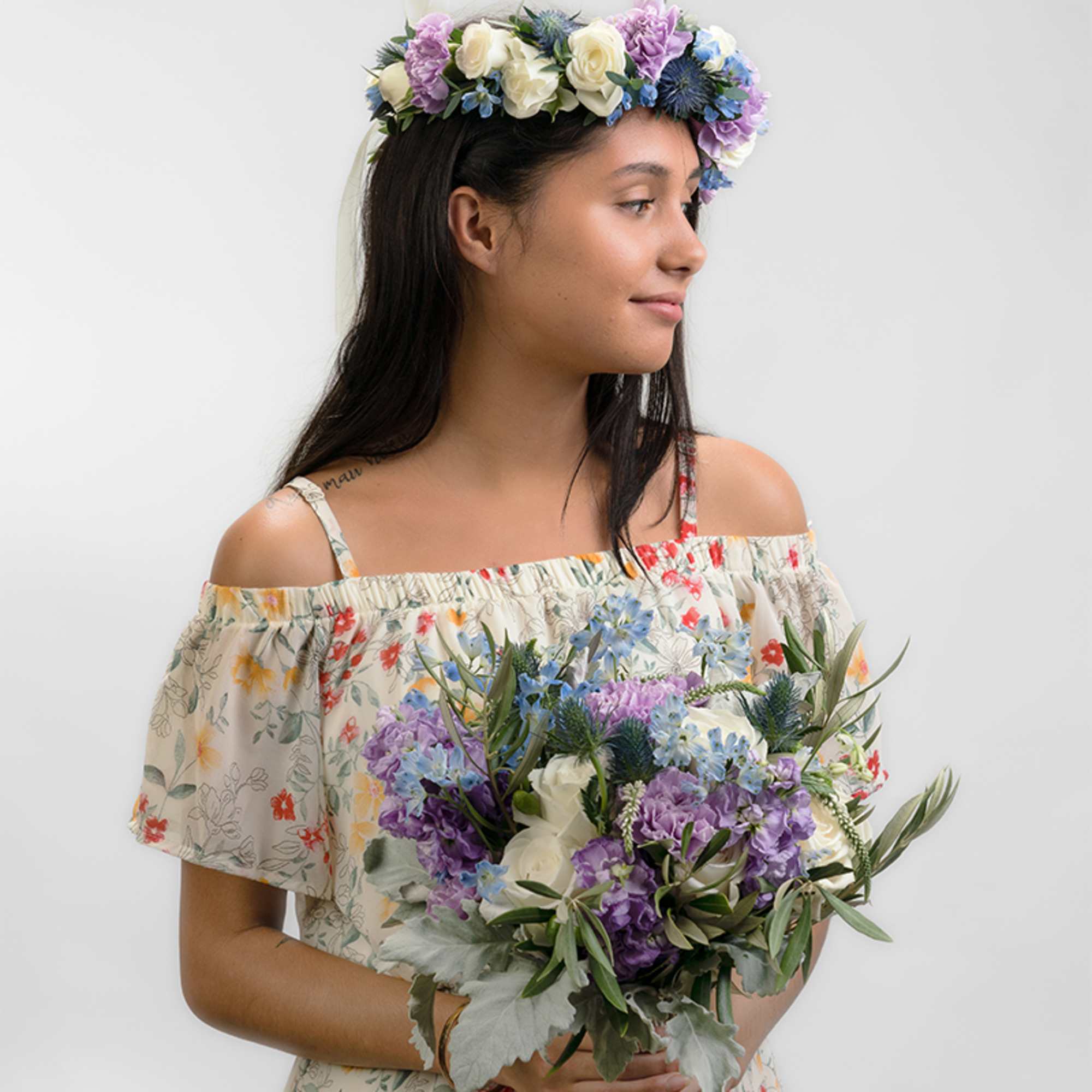 Young woman wearing a purple and white flower crown and holding a matching bouquet against a light background.
