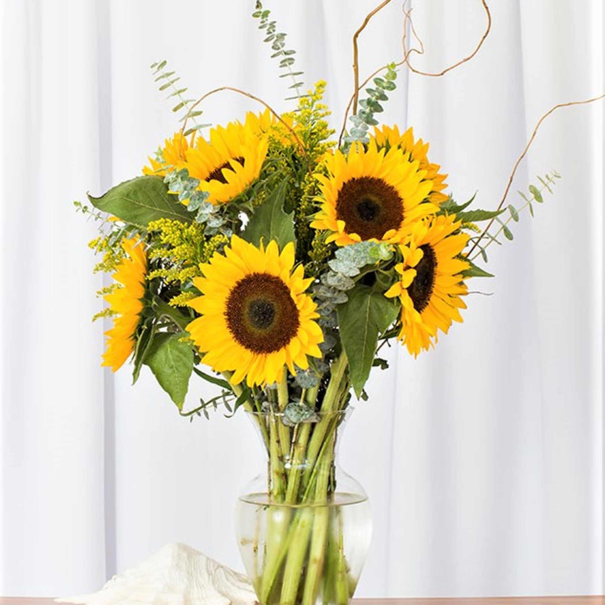 Tall bouquet of bright yellow sunflowers in a clear glass vase on a wooden surface.