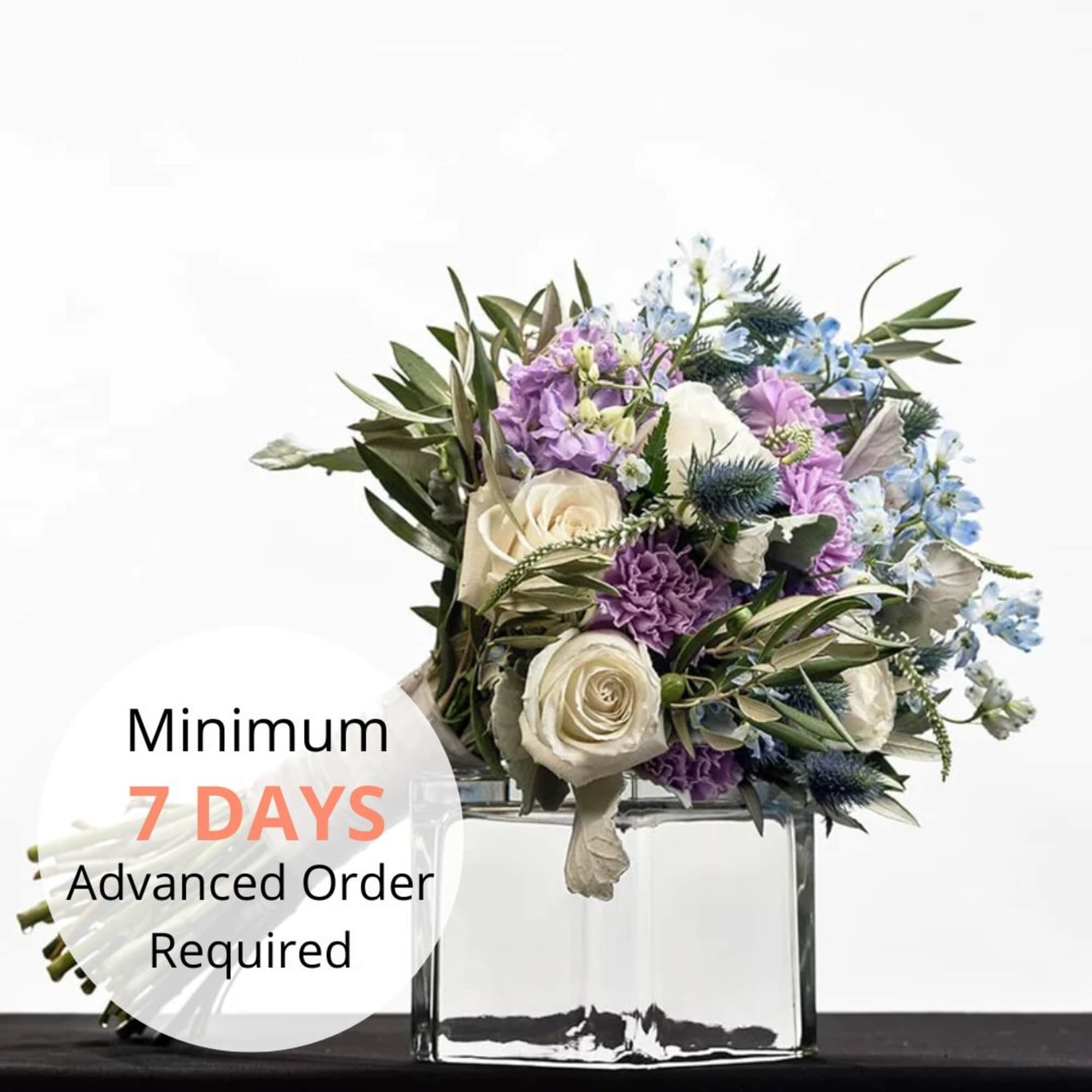 Hand-tied bouquet of ivory roses with purple and blue flowers in a clear glass cube vase