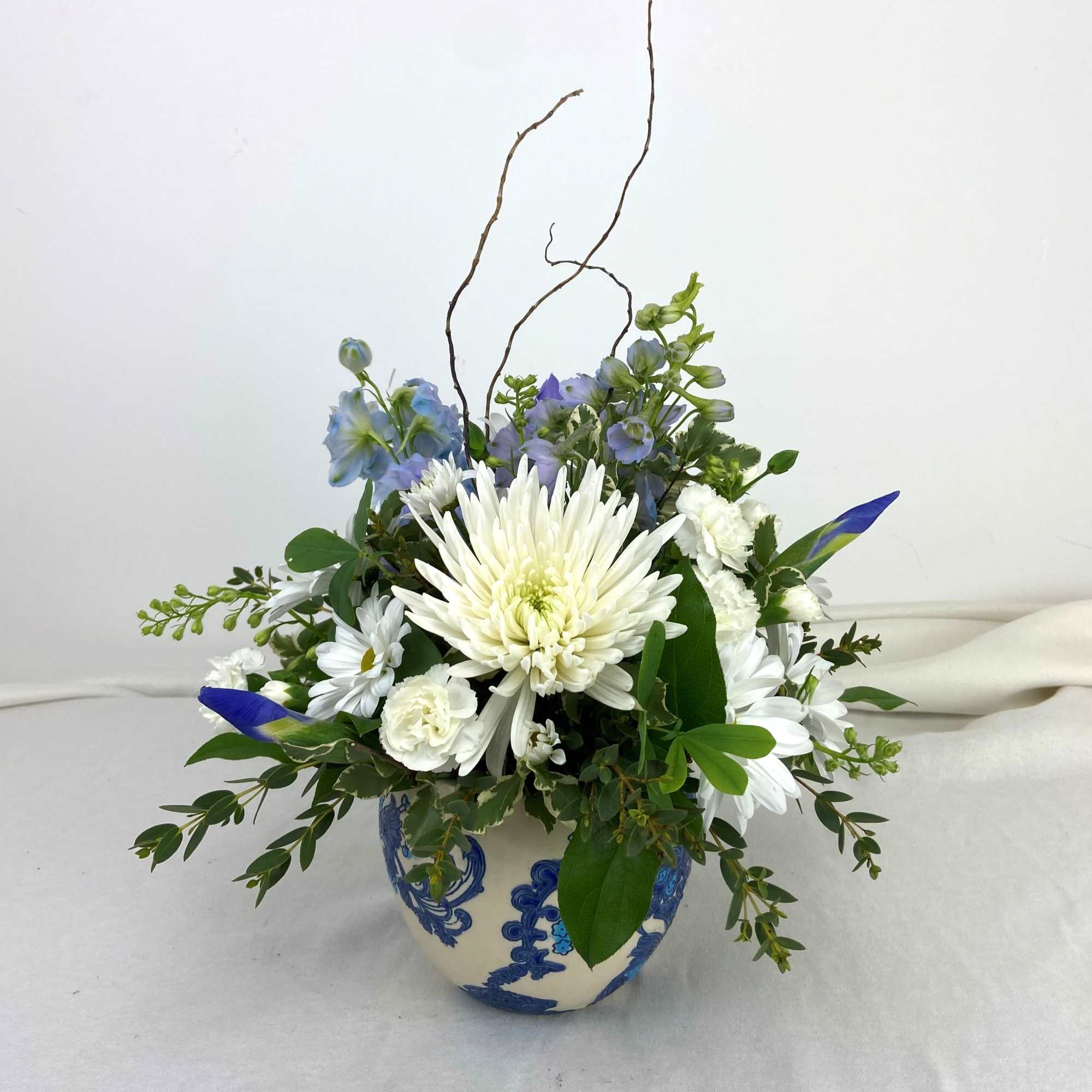 White chrysanthemums, pale blue delphinium and daisies arranged in a rounded 5.5"