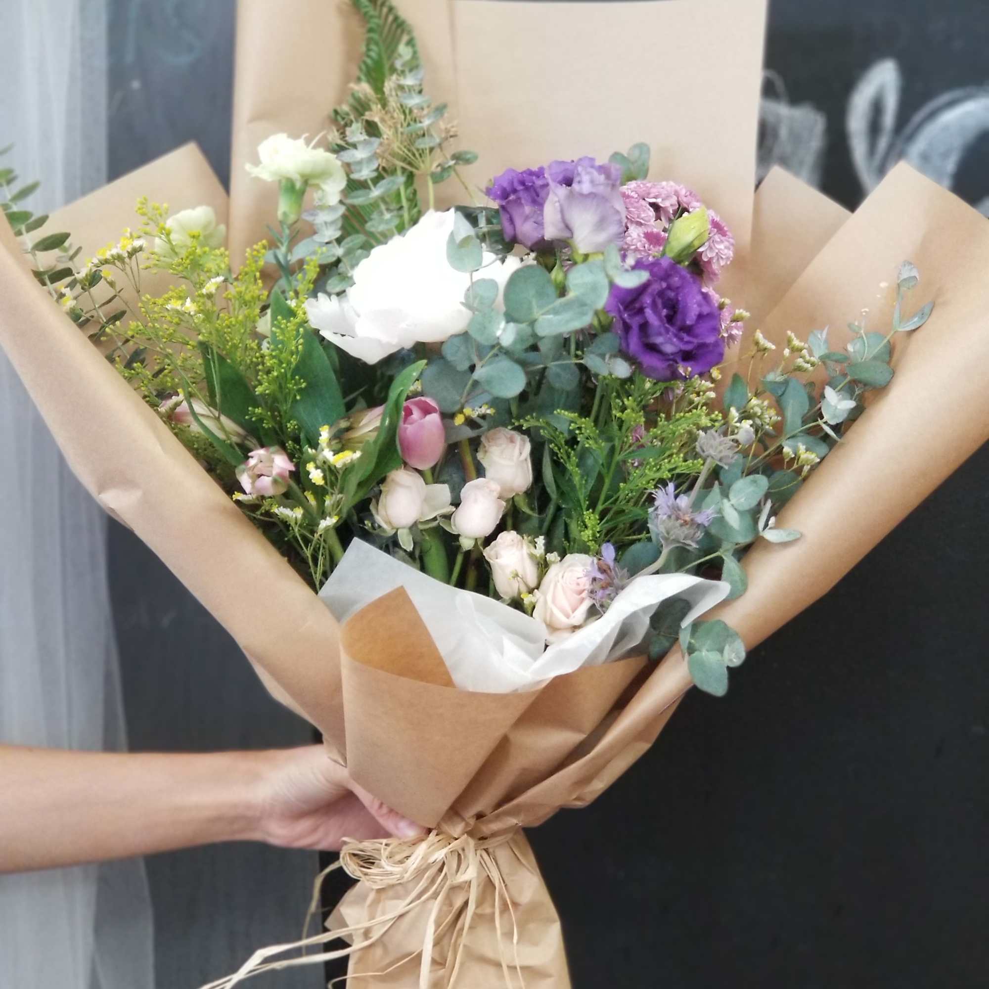 Hand-tied bouquet of purple, white, and pink flowers wrapped in kraft paper
