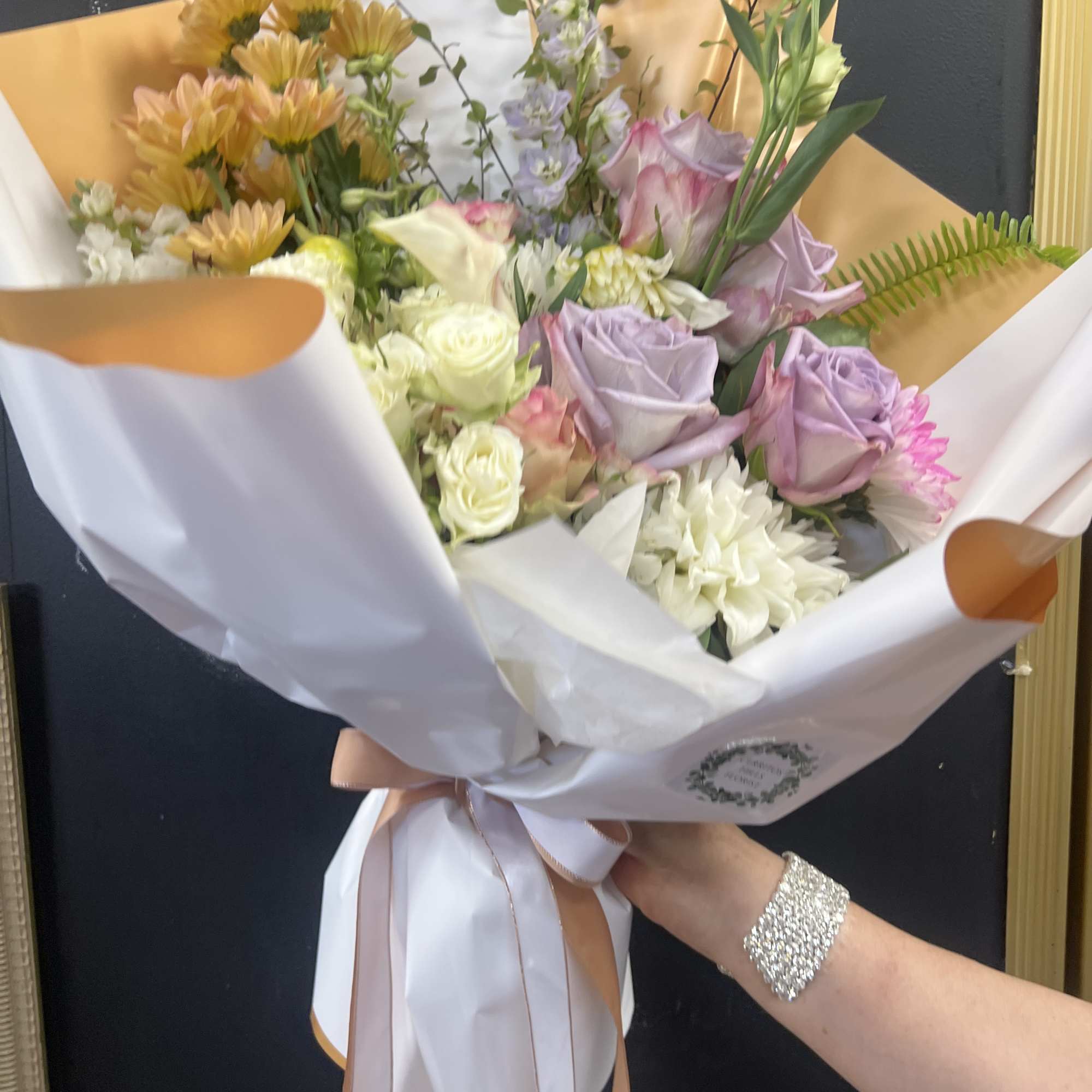 Handheld bouquet of pastel roses, chrysanthemums, and delphinium wrapped in white paper