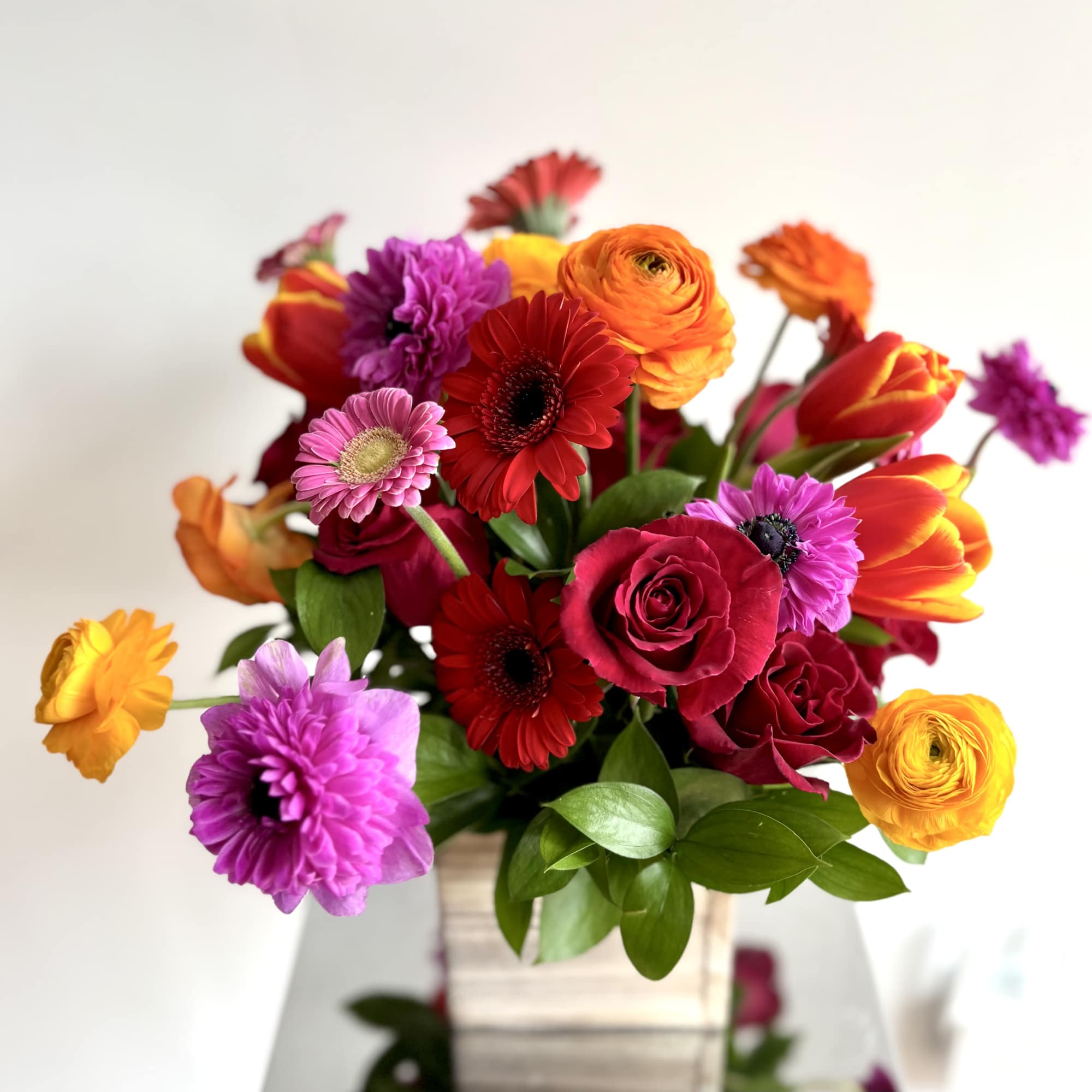 Bright mixed bouquet of gerbera daisies, roses, and ranunculus in a square vase