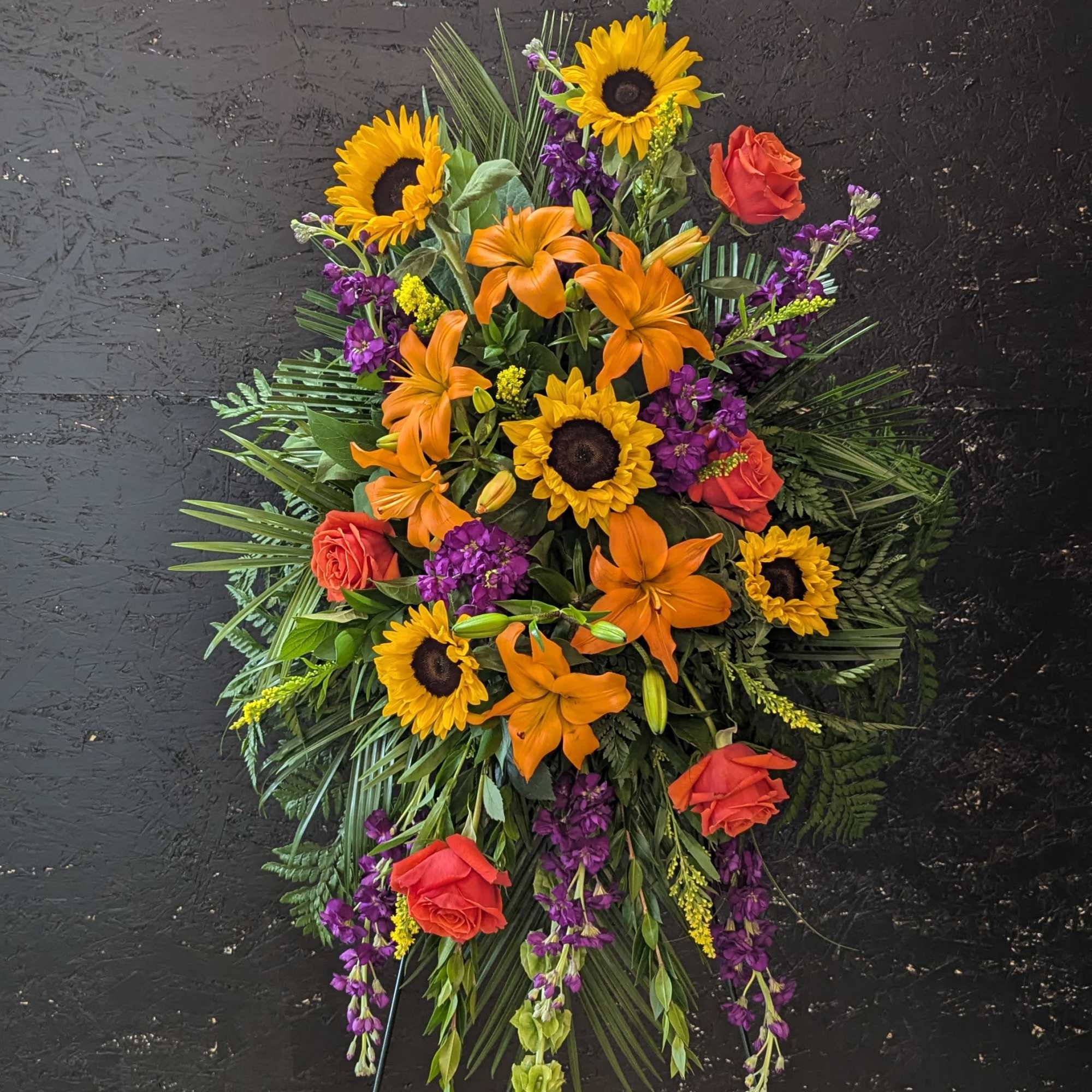 Standing spray includes, Orange Lilies, Sunflowers, Orange Roses, Violet Stock, Bells of