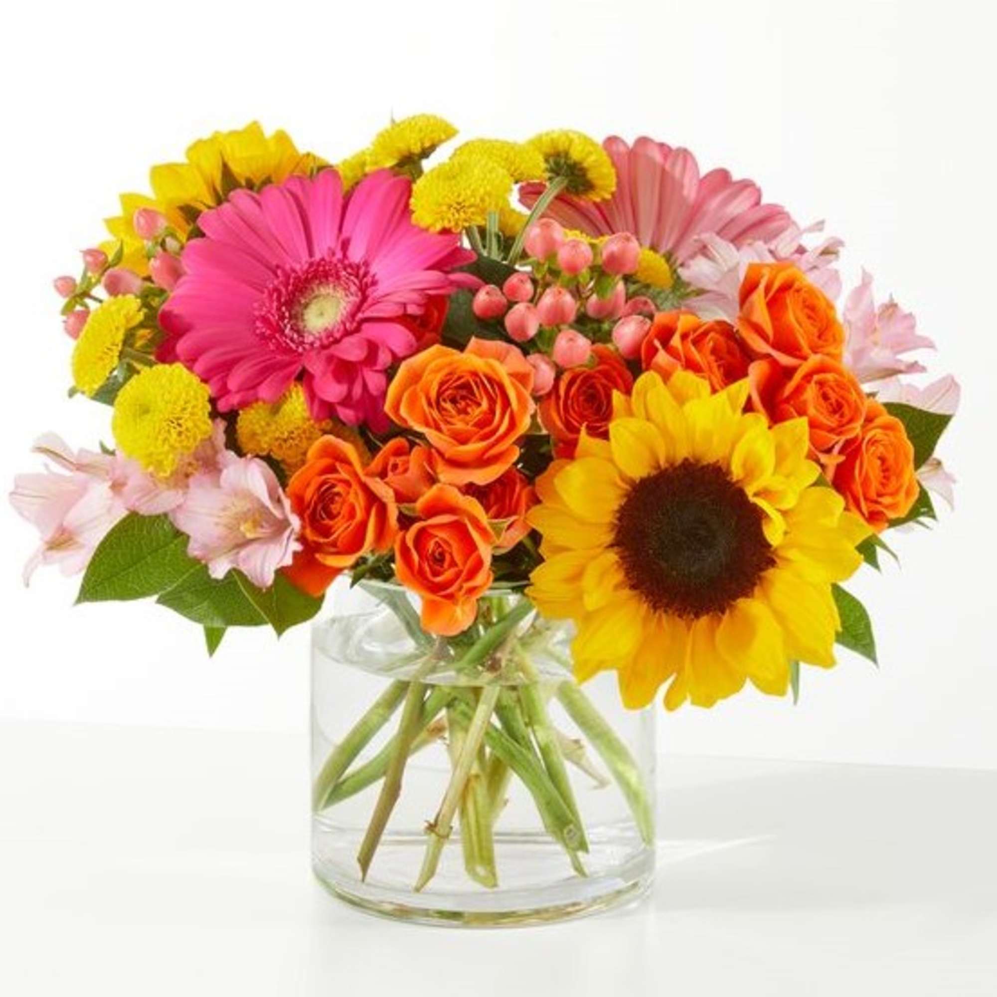 Mixed bouquet of gerbera daisies, roses, and sunflowers in a glass vase